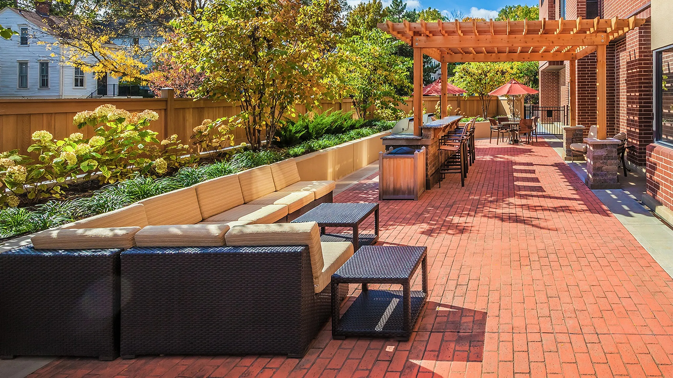 Inviting outdoor patio with wicker seating and brick flooring, featuring a shaded pergola and lush garden backdrop.