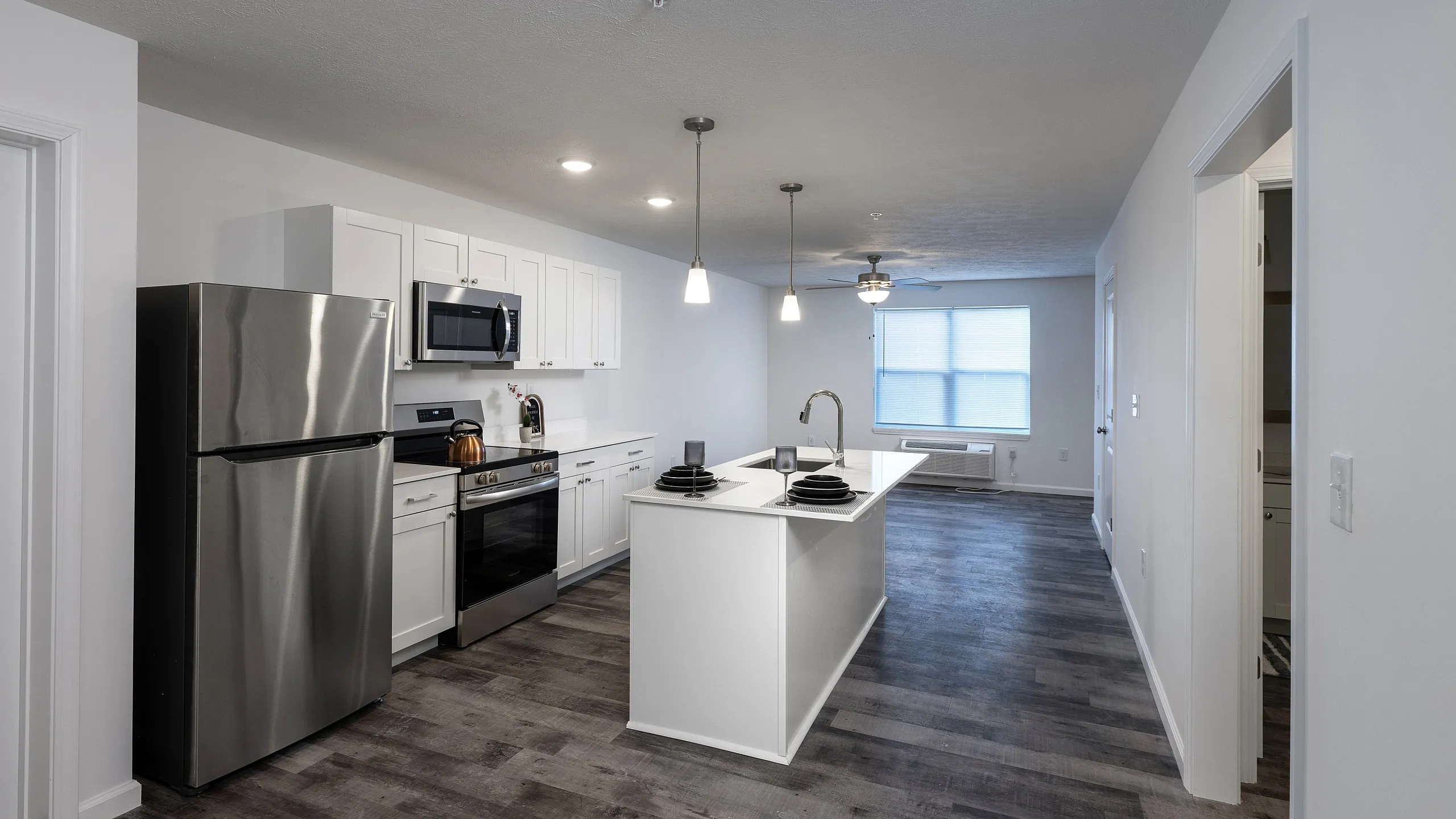 Modern apartment kitchen with sleek stainless steel appliances, an elegant island, and luxurious dark wood flooring leading to a spacious, sunlit living area.