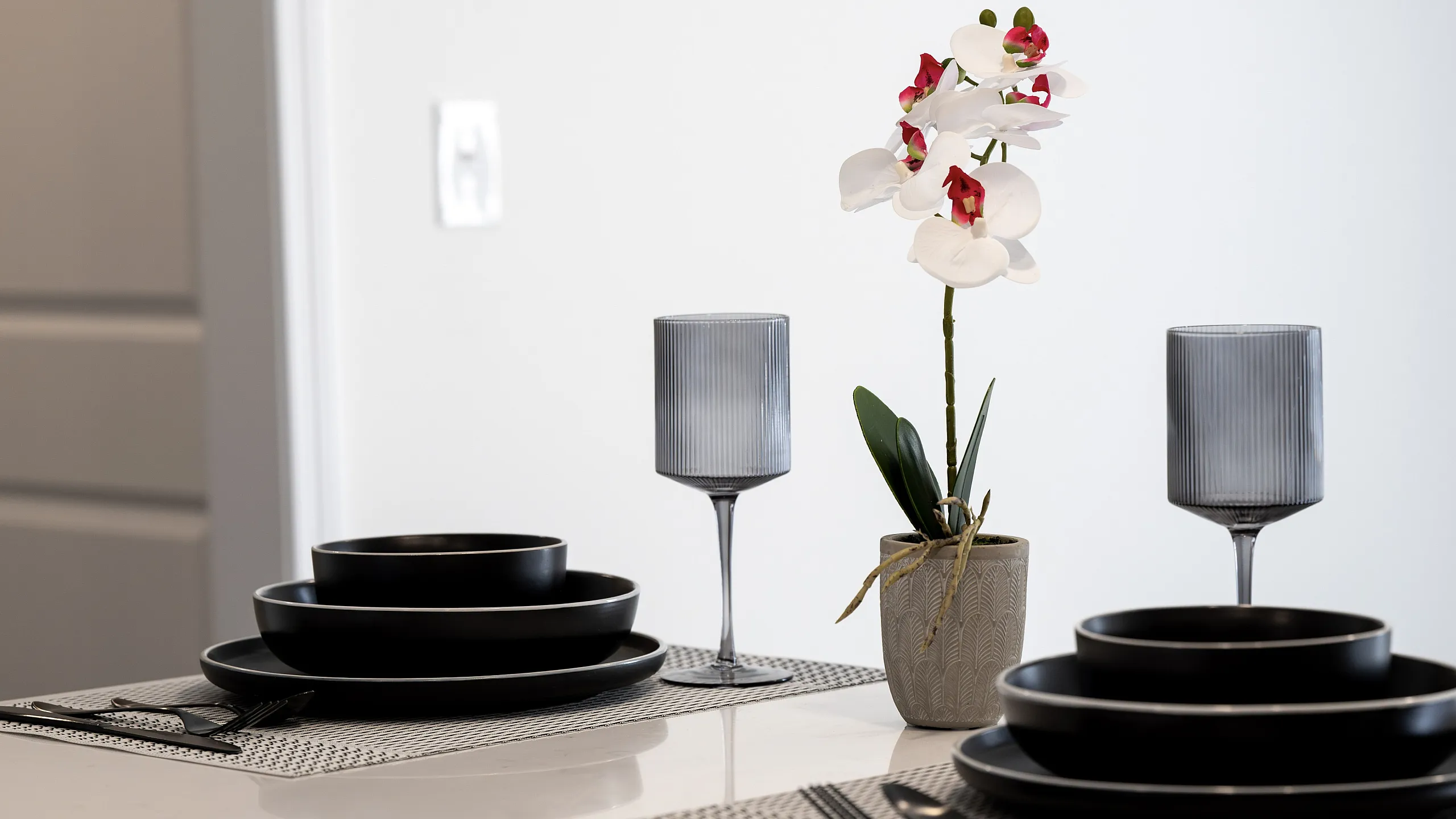Elegant dining setup featuring chic black dinnerware and delicate orchid centerpiece on a modern white table.