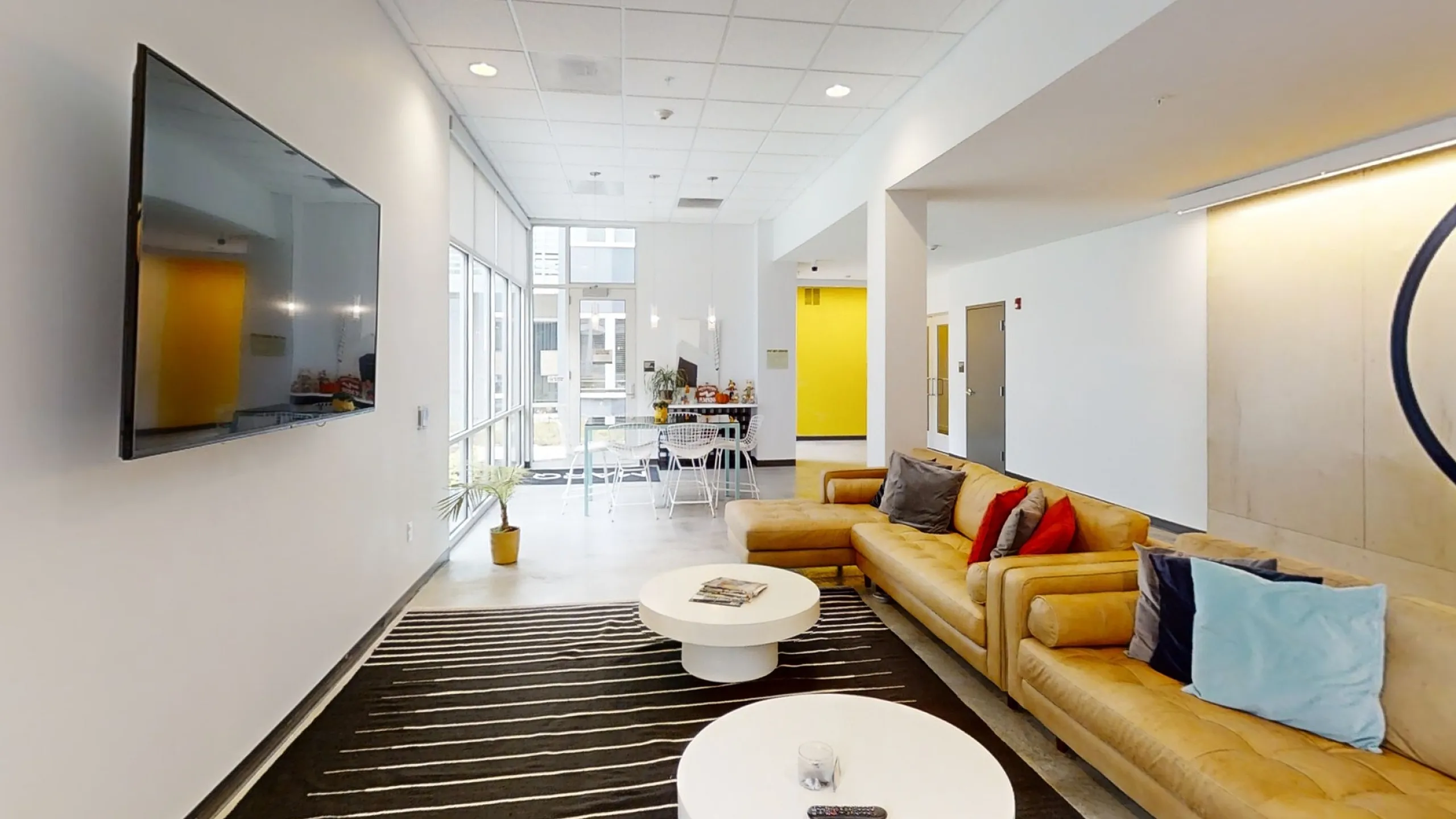 Modern apartment lounge with sleek flat-screen TV, cozy mustard-yellow sofa, vibrant decor, and stylish white tables, filled with natural light from expansive windows.
