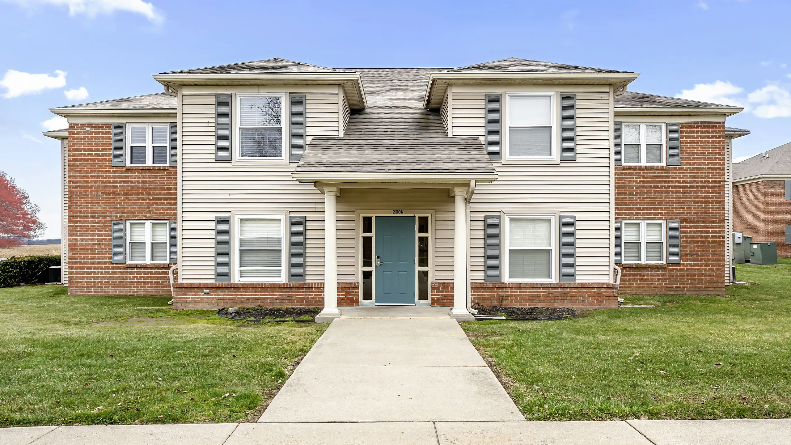 Charming two-story brick and siding apartment building with large windows and a welcoming entrance, surrounded by well-maintained green lawns under a clear blue sky.
