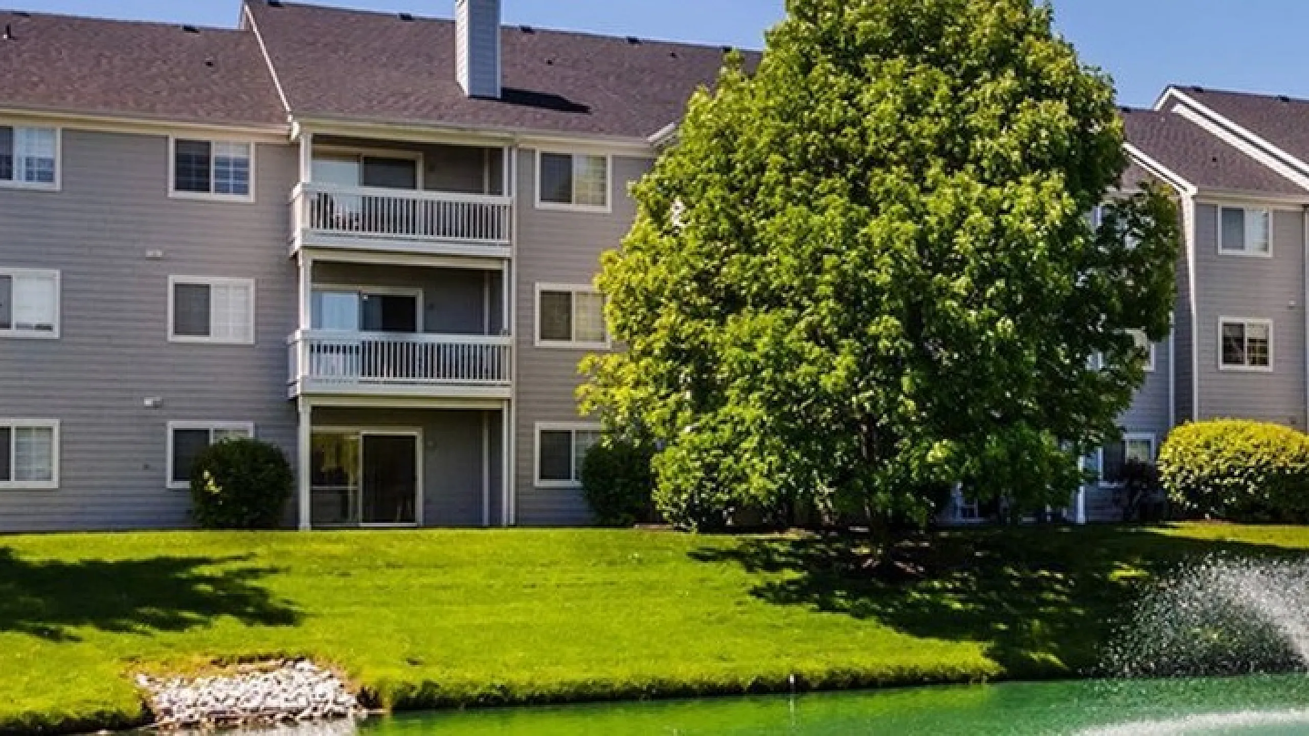 Charming apartment complex with serene pond and fountain, surrounded by lush greenery and vibrant landscape.