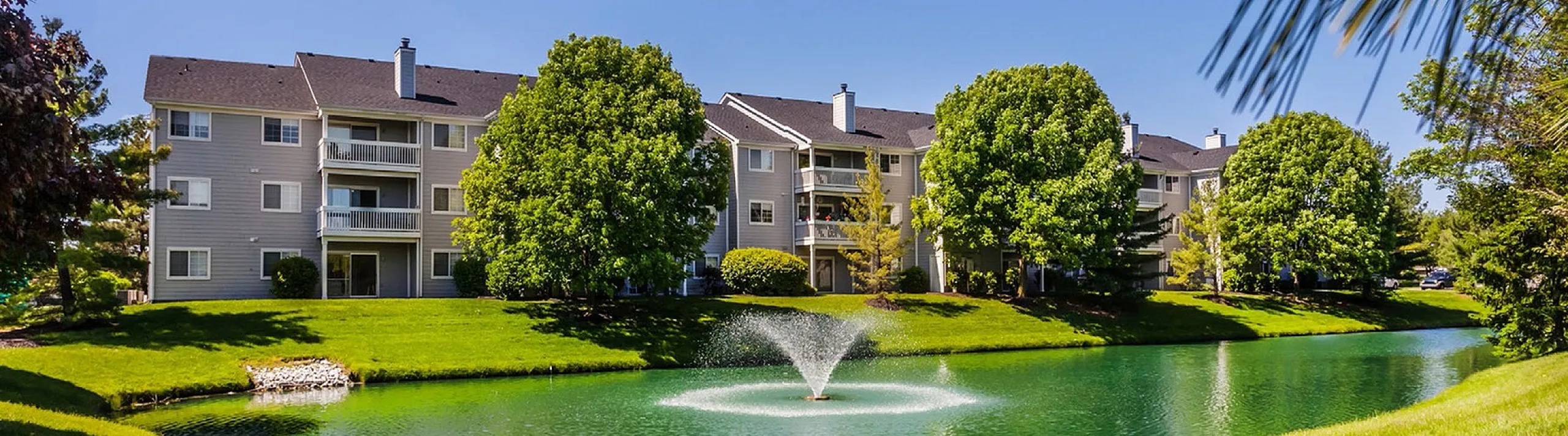 Charming apartment complex set amongst lush greenery, featuring a serene pond and fountain creating a picturesque living environment.