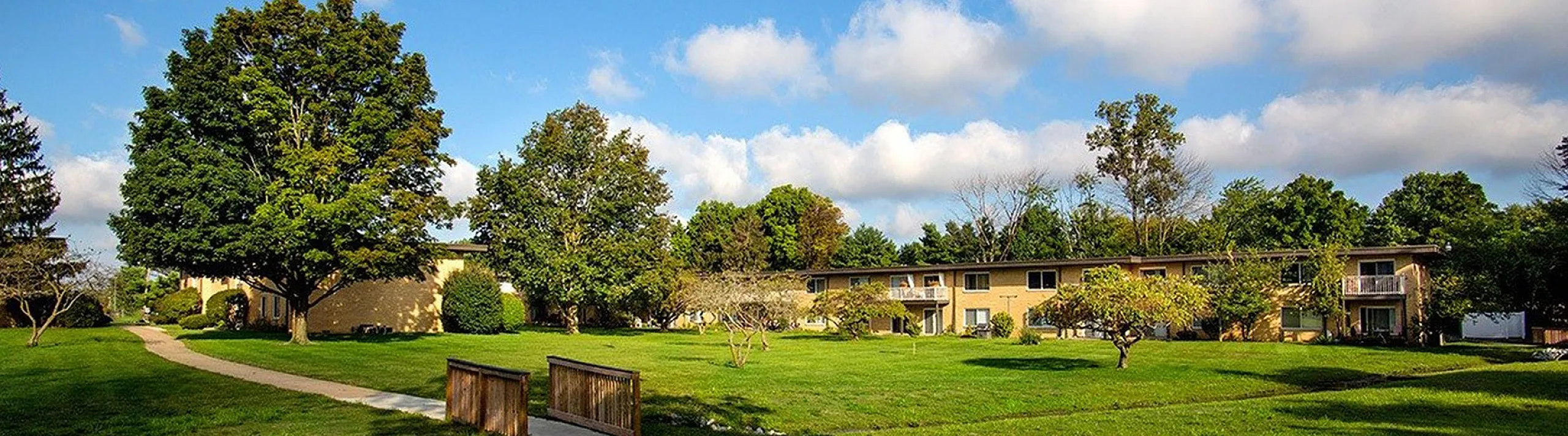Charming apartment complex surrounded by lush green lawns and mature trees under a clear blue sky.