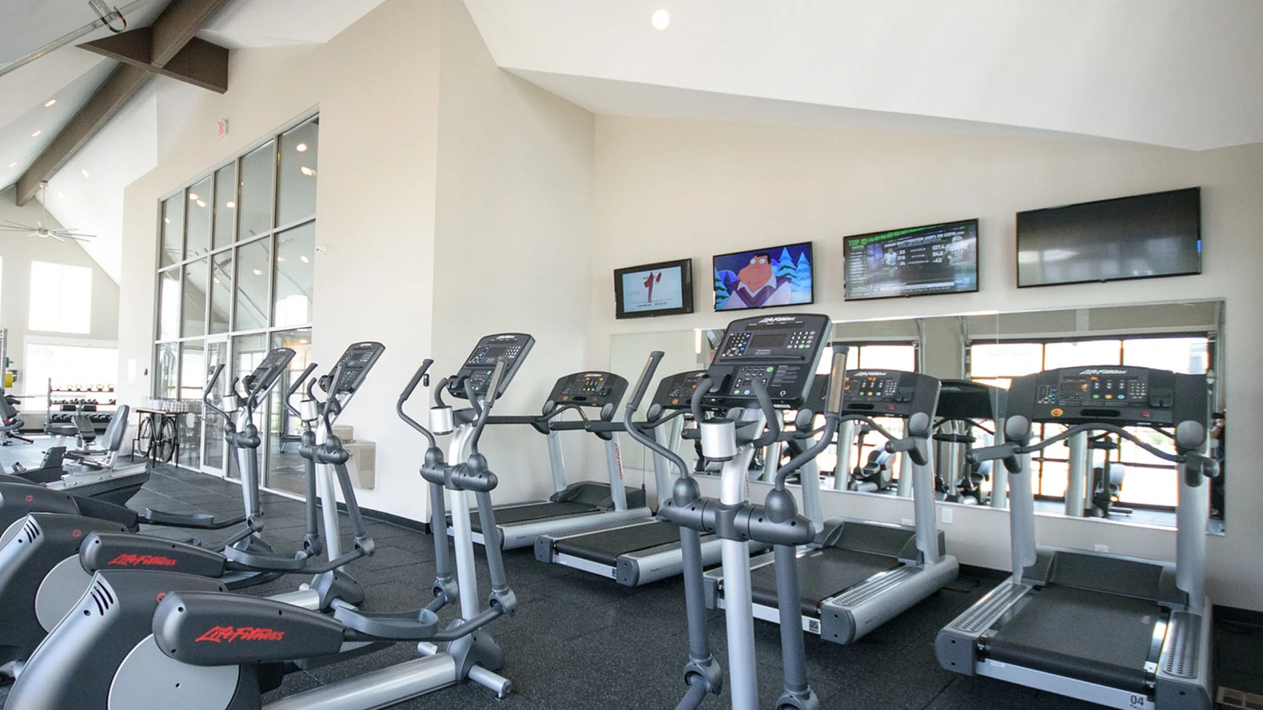 Modern fitness center featuring state-of-the-art ellipticals and treadmills with wall-mounted TVs and ample natural light.