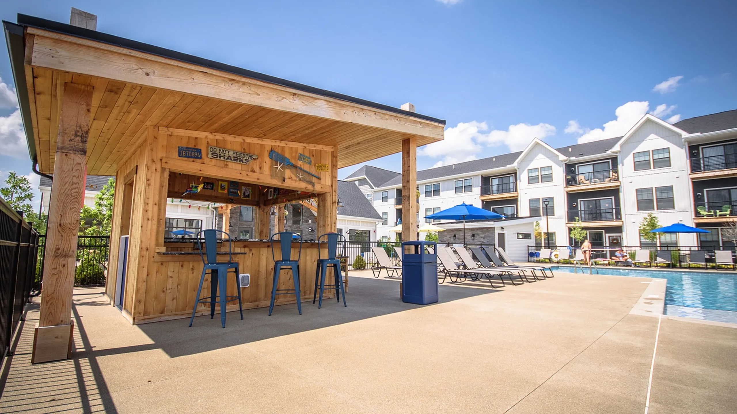 Vibrant apartment complex featuring a charming wooden poolside bar, surrounded by stylish lounge chairs and a sparkling swimming pool under clear blue skies.