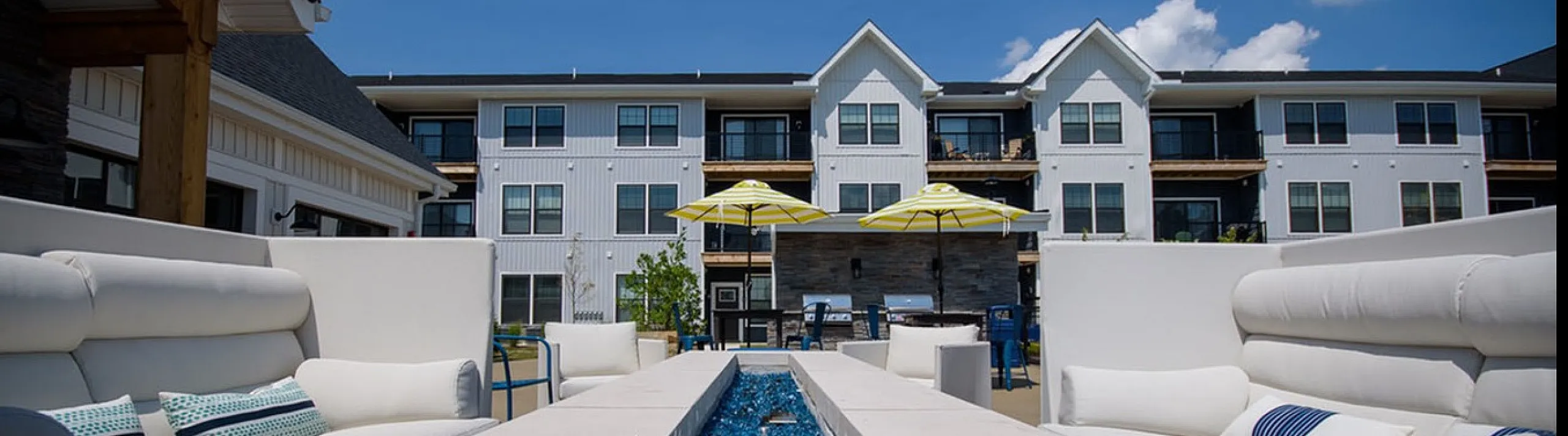 Luxurious apartment exterior with modern white facade, featuring a stunning outdoor lounge with cozy seating and elegant blue fire pit under a clear blue sky.