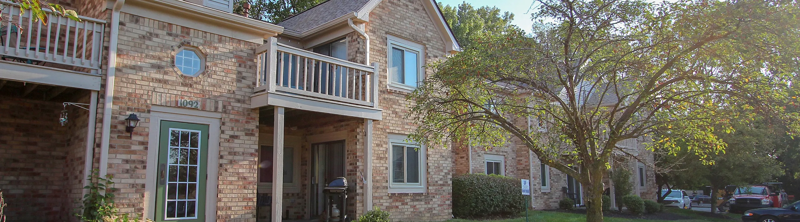 Charming brick apartment building surrounded by lush greenery, featuring a welcoming front porch and sunlit balconies.