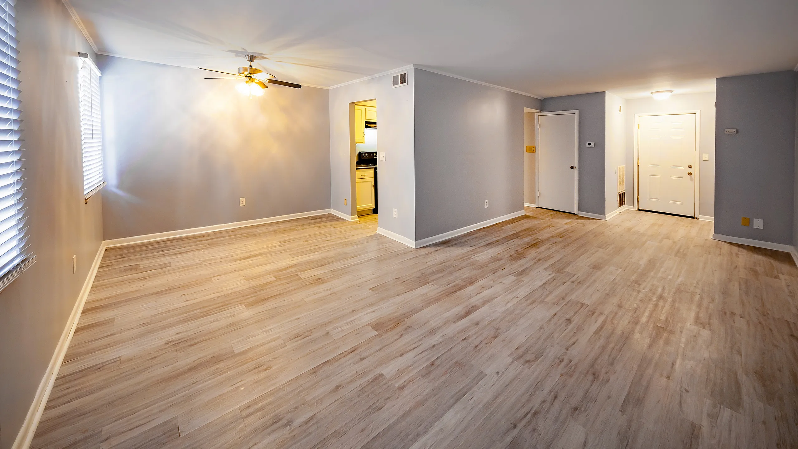 Spacious, well-lit living room with elegant wooden flooring and modern ceiling fan, leading to an inviting kitchen.