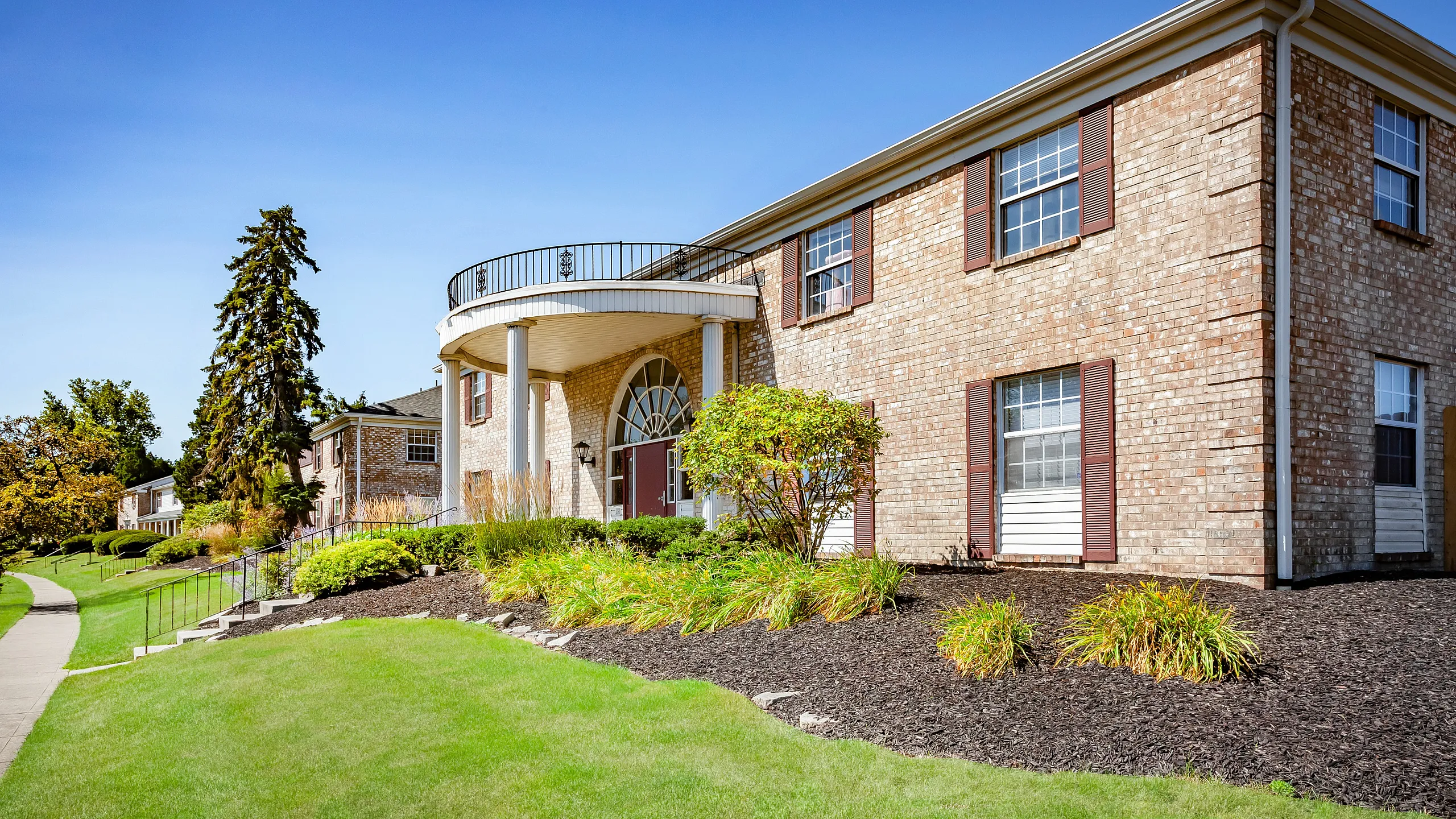 Charming two-story brick apartment building with elegant arches and lush green landscaping under a clear blue sky.