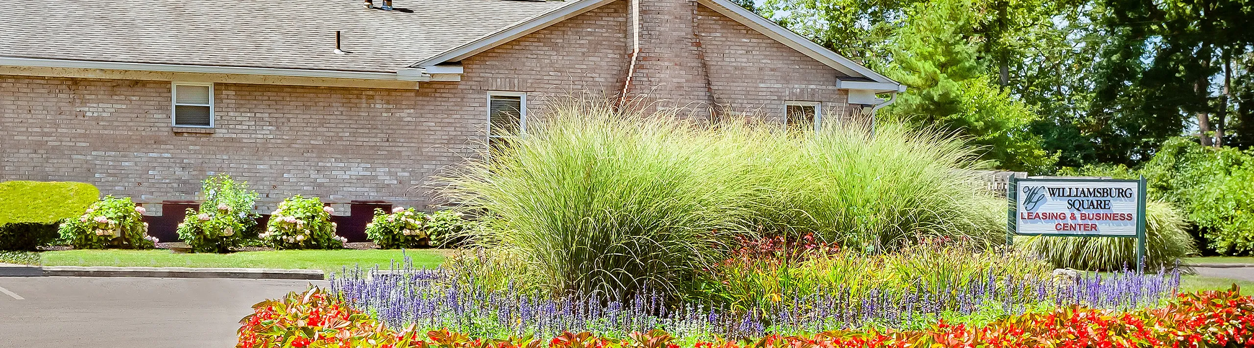 Inviting brick building surrounded by vibrant flower beds and lush greenery under a bright blue sky, featuring 'Williamsburg Square Leasing & Business Center' sign.