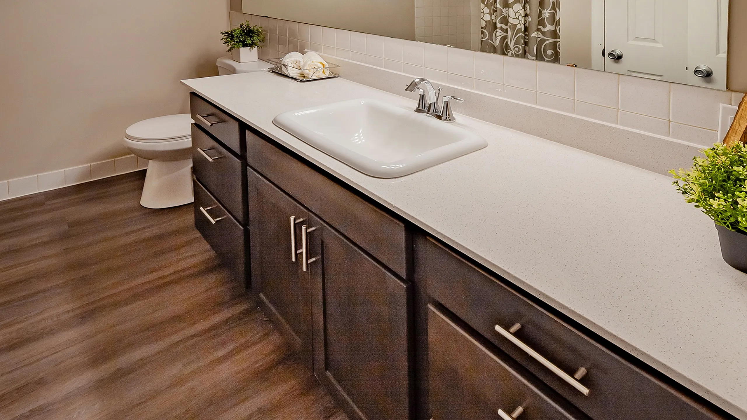 Elegant bathroom with dark wood cabinets, sleek countertop, and large mirror, featuring modern lighting and lush greenery.