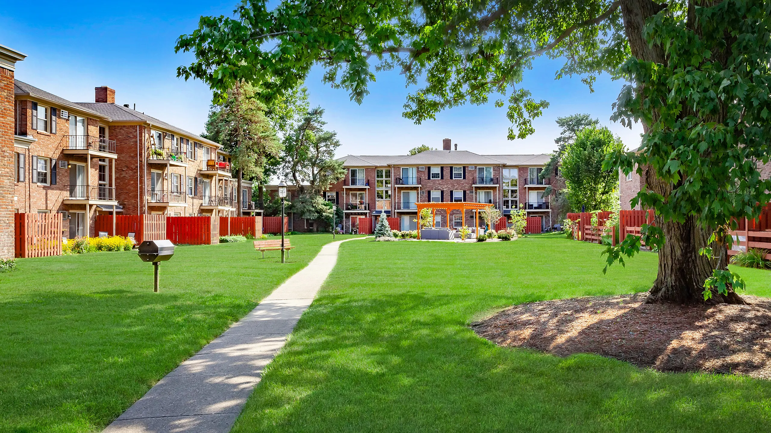 Charming apartment complex with brick exterior set in a lush green courtyard, featuring a beautiful outdoor seating area under a wooden arbor.