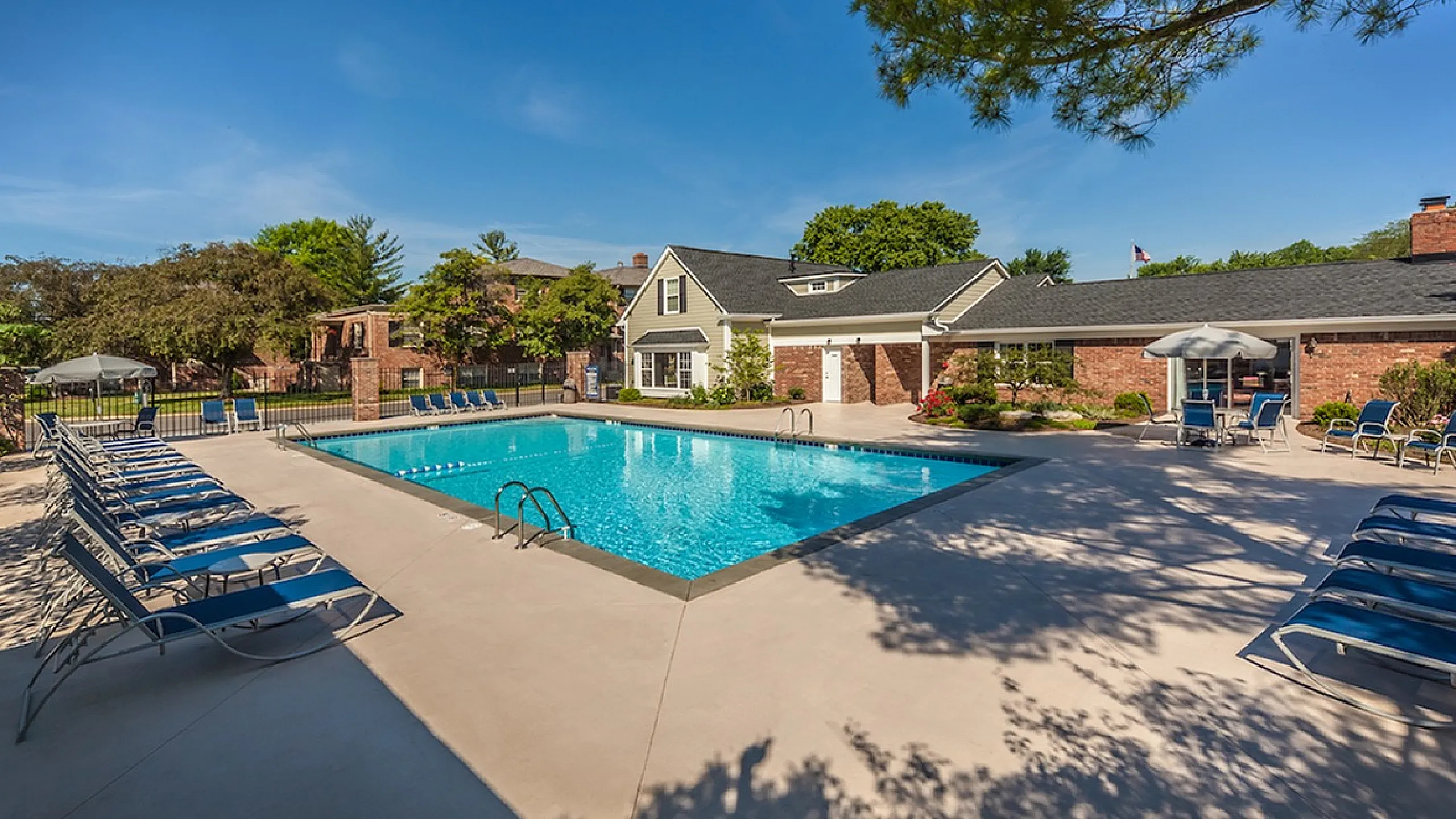 Inviting outdoor swimming pool area with lounge chairs and umbrellas, surrounded by lush greenery and a charming brick clubhouse.