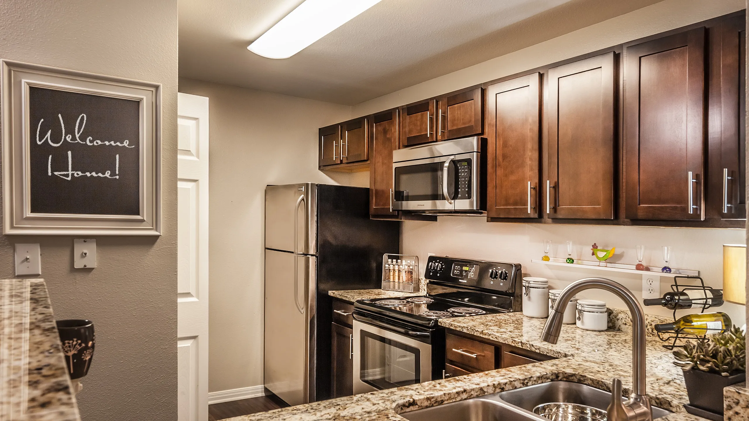 Modern kitchen with granite countertops, stainless steel appliances, and elegant cabinetry, featuring a welcoming sign on the wall.
