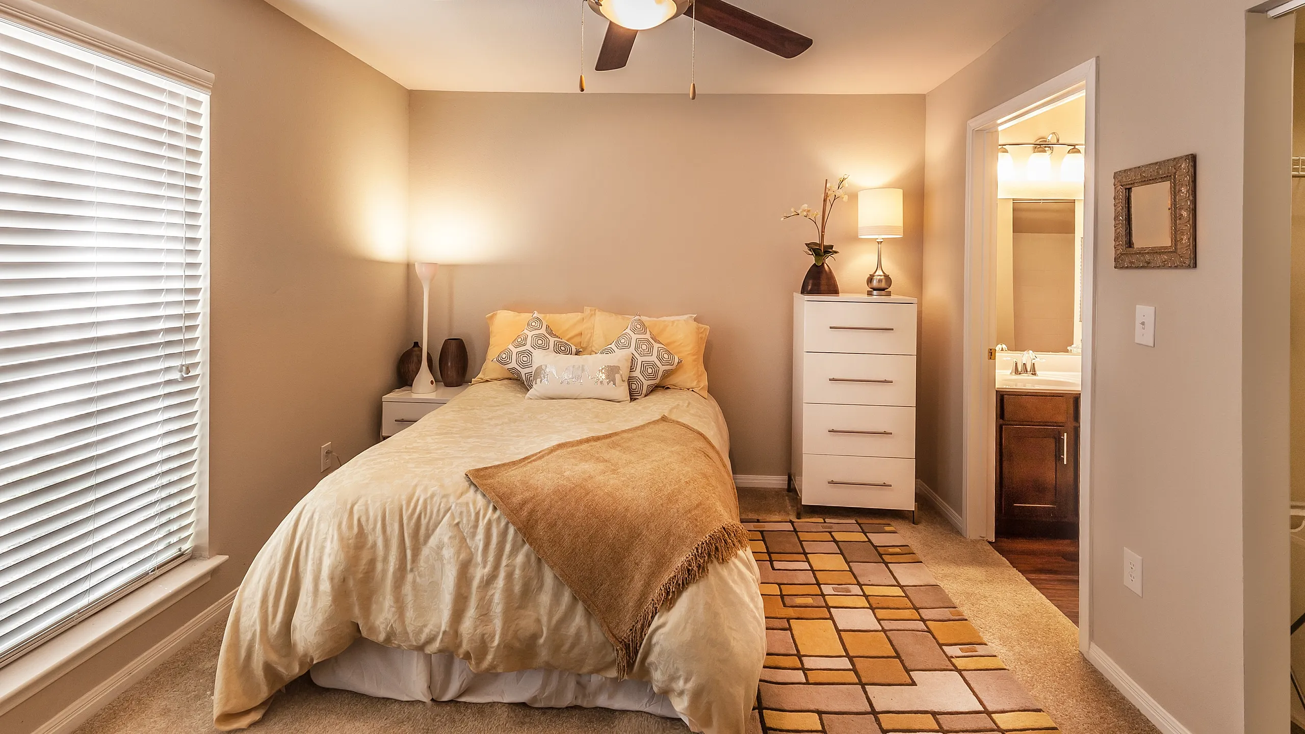 Inviting bedroom with cozy bedding, warm lighting, and elegant decor, featuring a modern ceiling fan and soft carpet flooring.