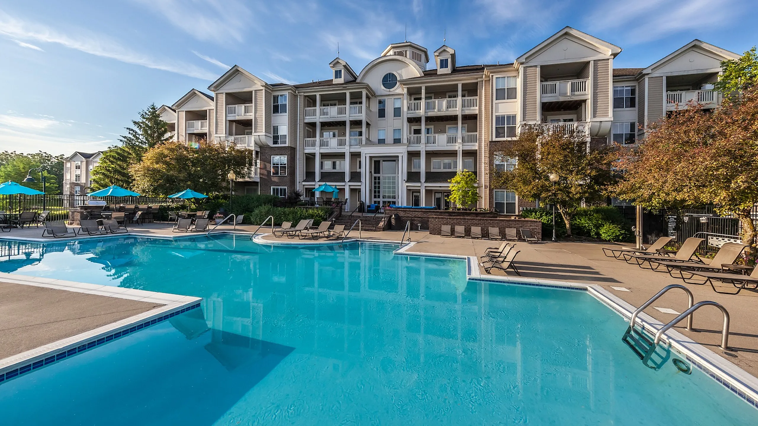 Luxurious apartment complex with a stunning outdoor pool surrounded by sun loungers and lush greenery, set against a clear blue sky.