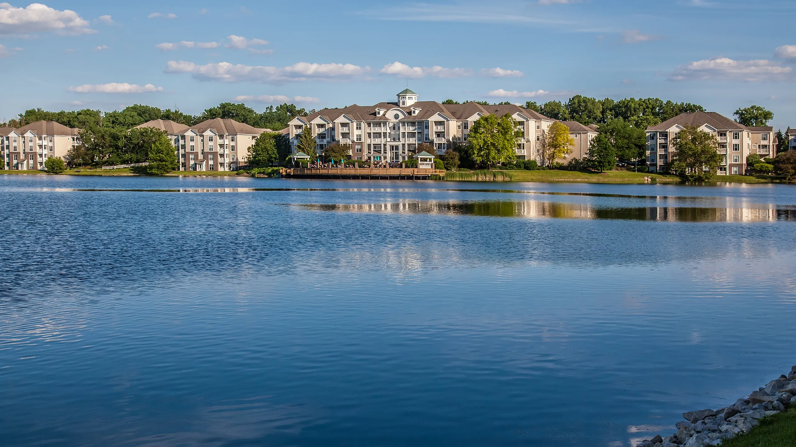 Scenic lakeside apartment complex with elegant architecture amidst lush greenery and a tranquil water view.