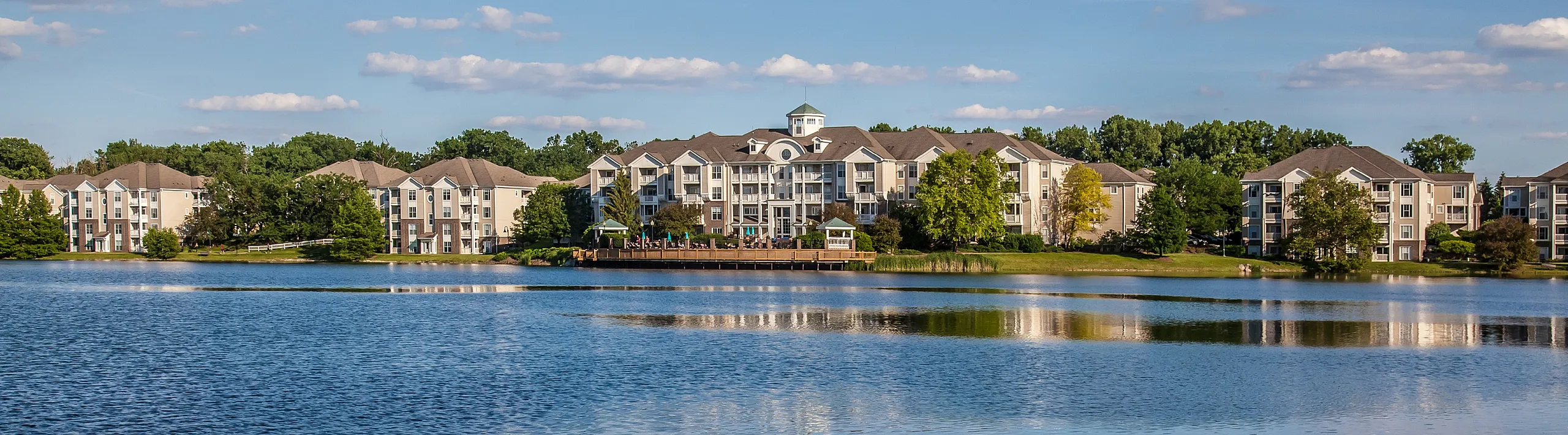 Elegant apartment complex set against a serene lakeside backdrop, featuring lush greenery and beautiful reflections of modern architecture on the water.