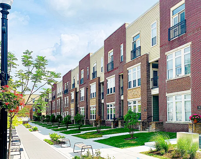 Modern row of townhouses with charming brick facades and inviting balconies, adjacent to a well-maintained pedestrian walkway lined with benches and greenery.
