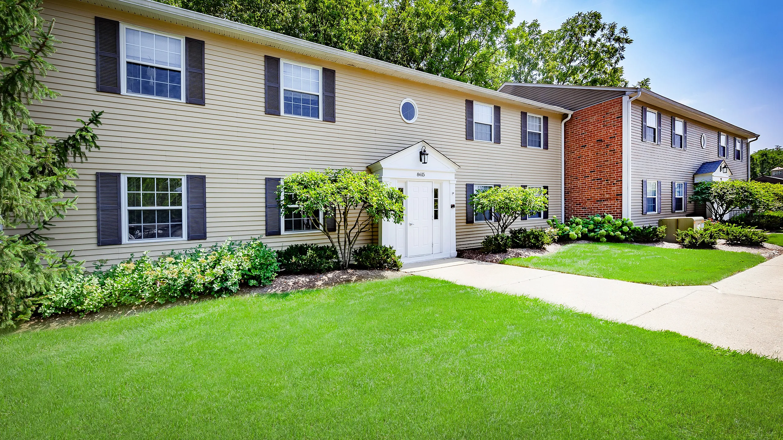 Charming two-story apartment complex with lush landscaping, vibrant greenery, and classic brick and siding exterior.