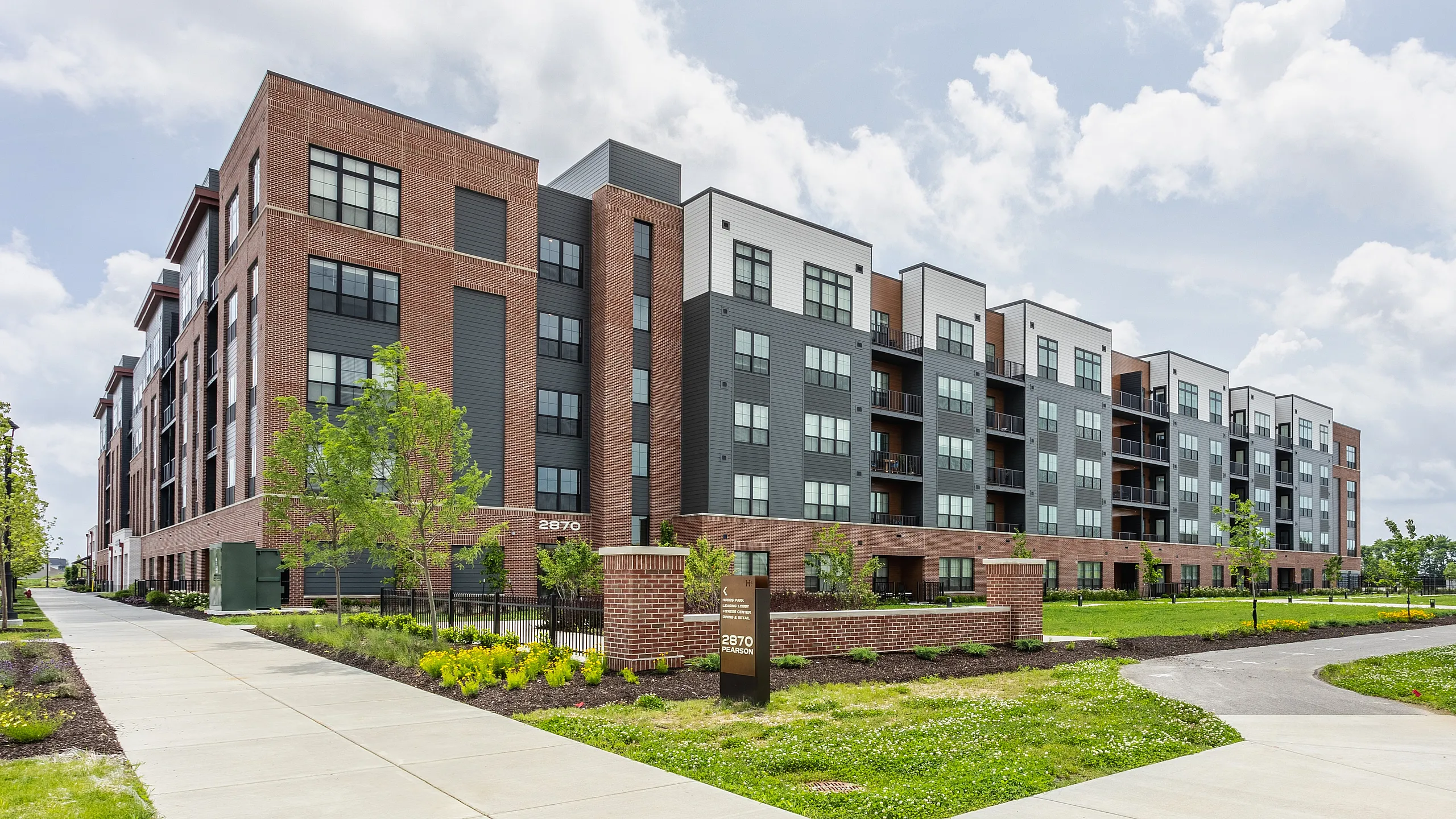 Modern multi-story apartment complex with stylish brick and panel exterior, surrounded by manicured gardens and pathways.