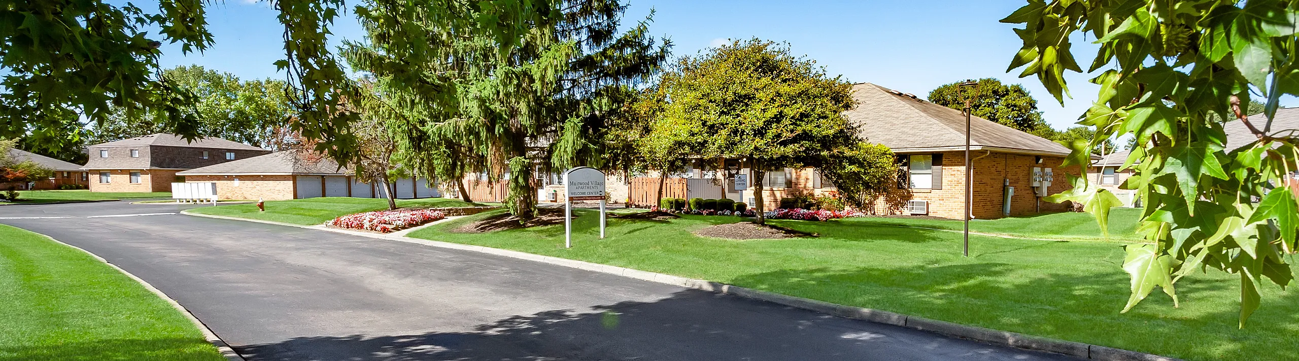 Charming brick apartment home surrounded by lush green lawns and mature trees on a sunny day, offering serene community living.
