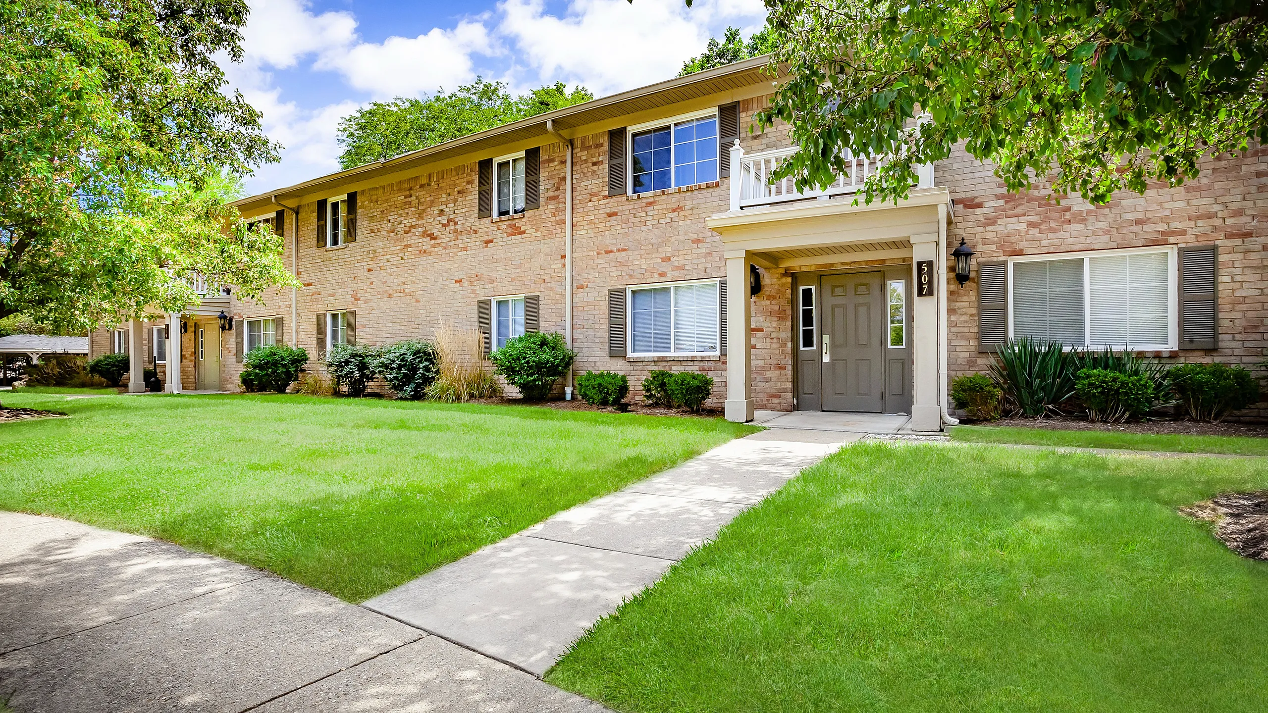 Charming apartment exterior with lush green lawn and inviting entrance, surrounded by mature trees and vibrant landscaping.