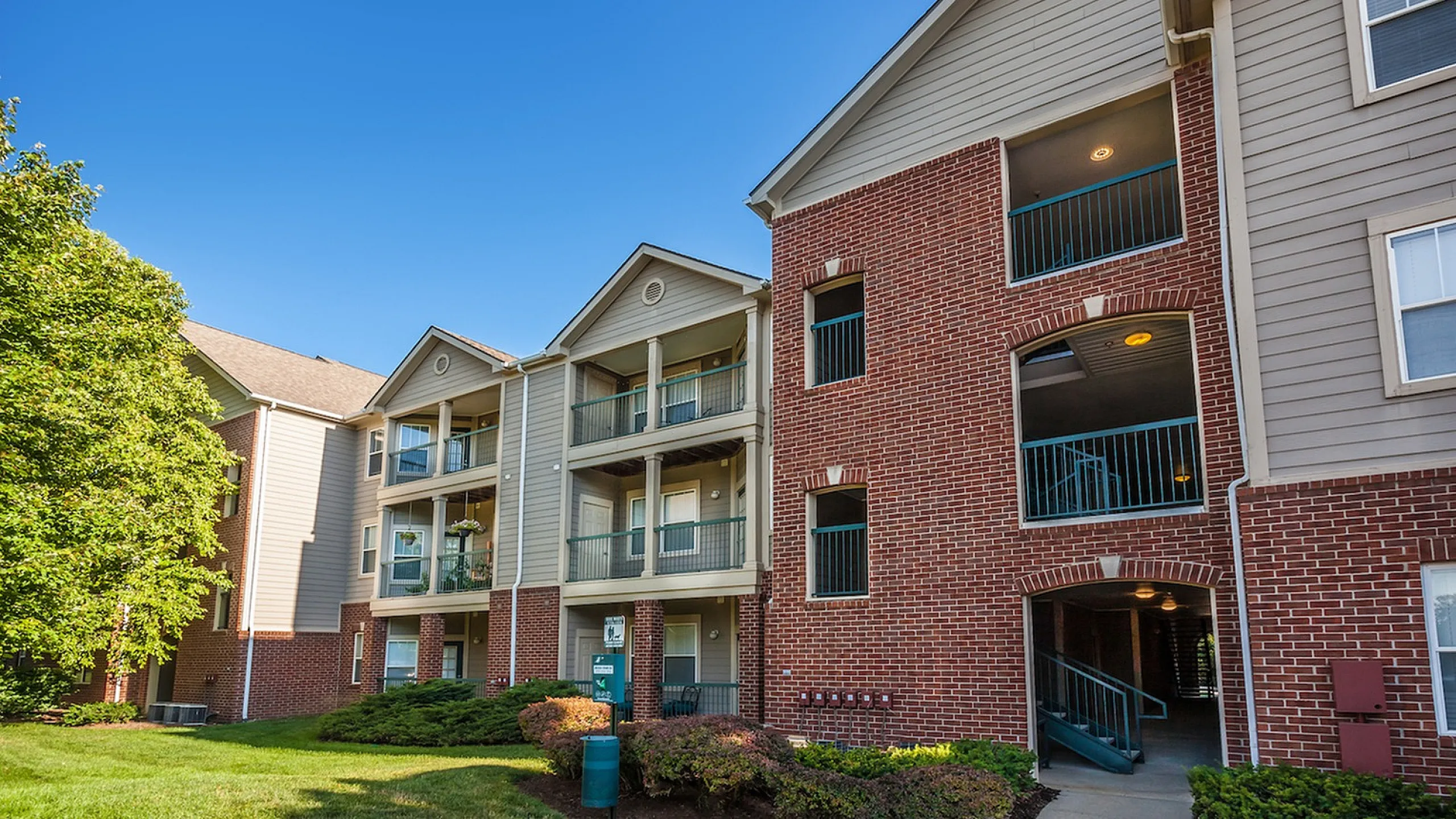 Charming apartment building with red brick facade, spacious balconies, and lush green surroundings under a bright blue sky.