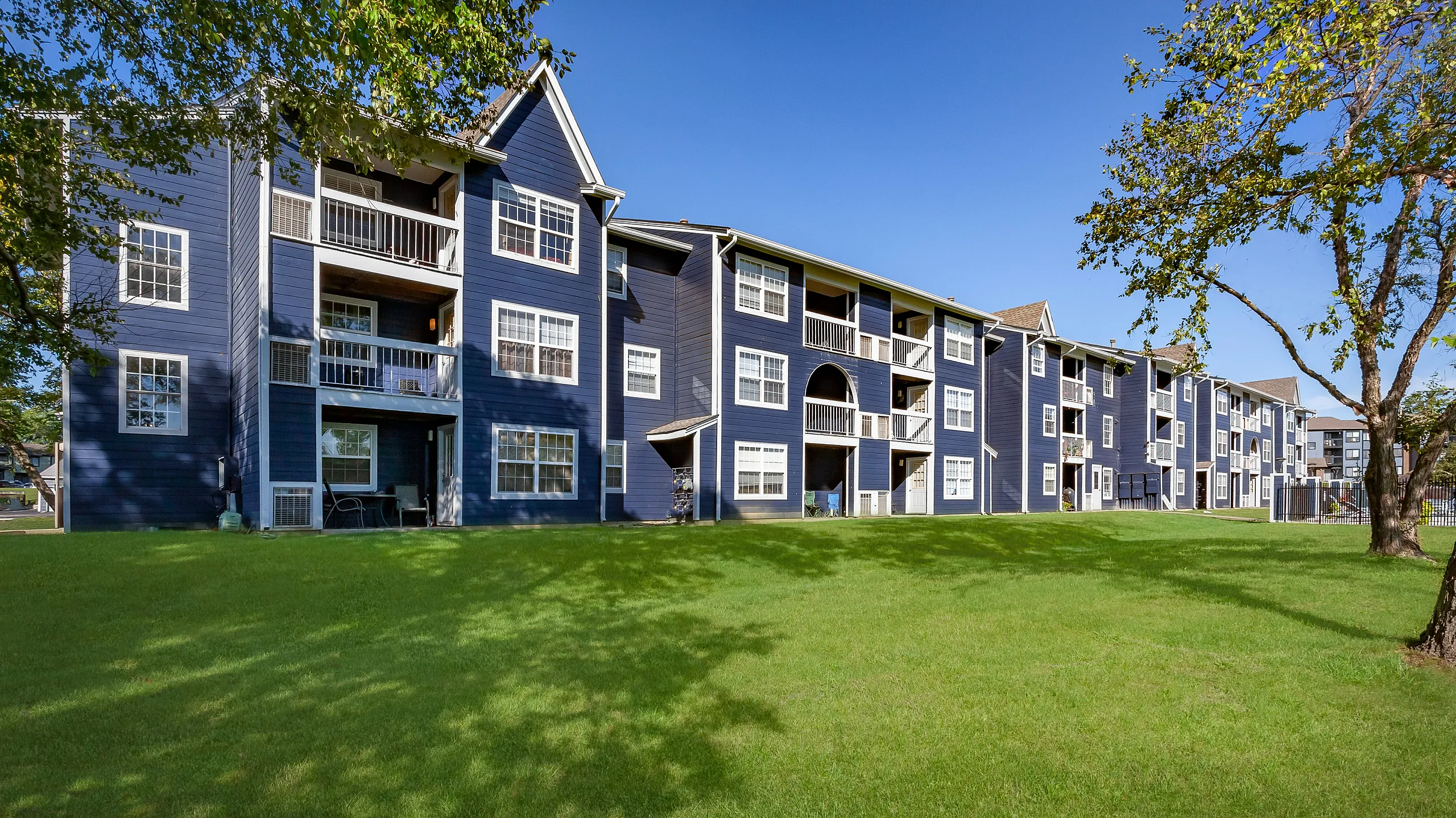 Charming blue apartment complex with spacious balconies, nestled in lush greenery under a clear blue sky.