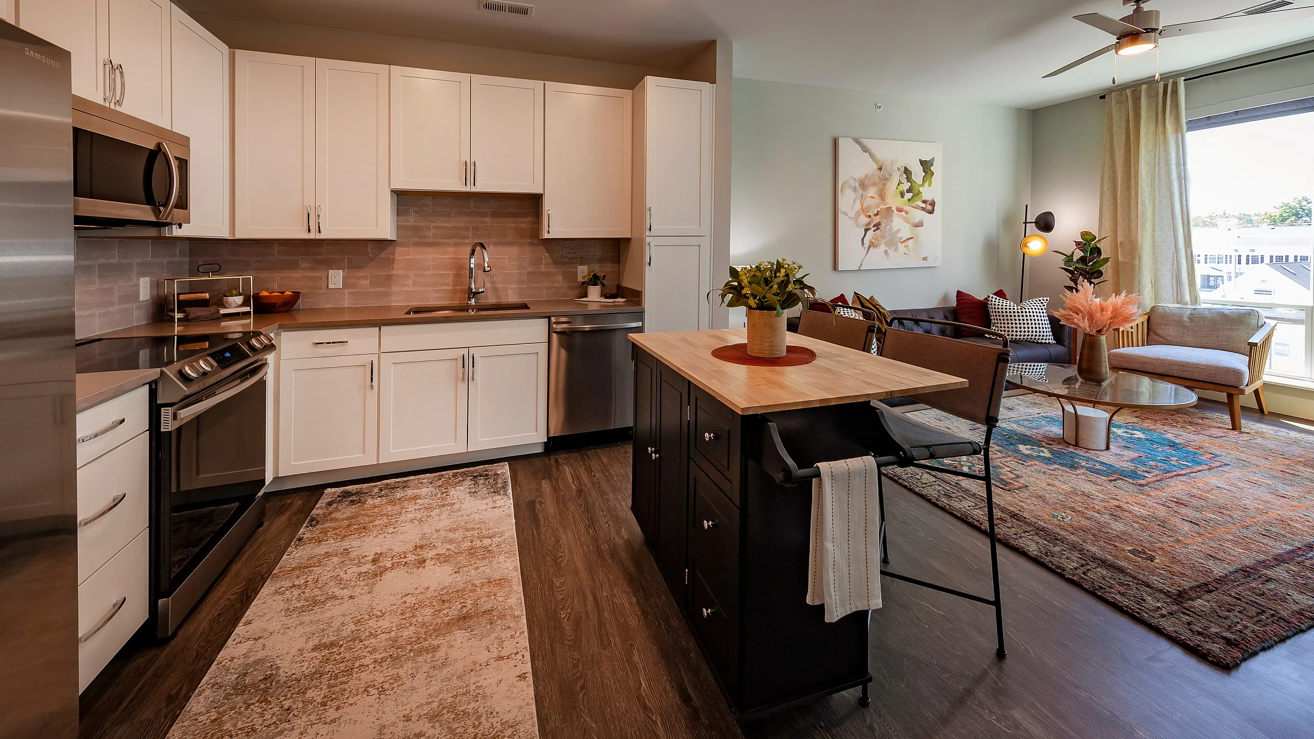 Modern apartment interior showcasing a sleek kitchen with white cabinets, stainless steel appliances, and a spacious living area with stylish decor and large windows.