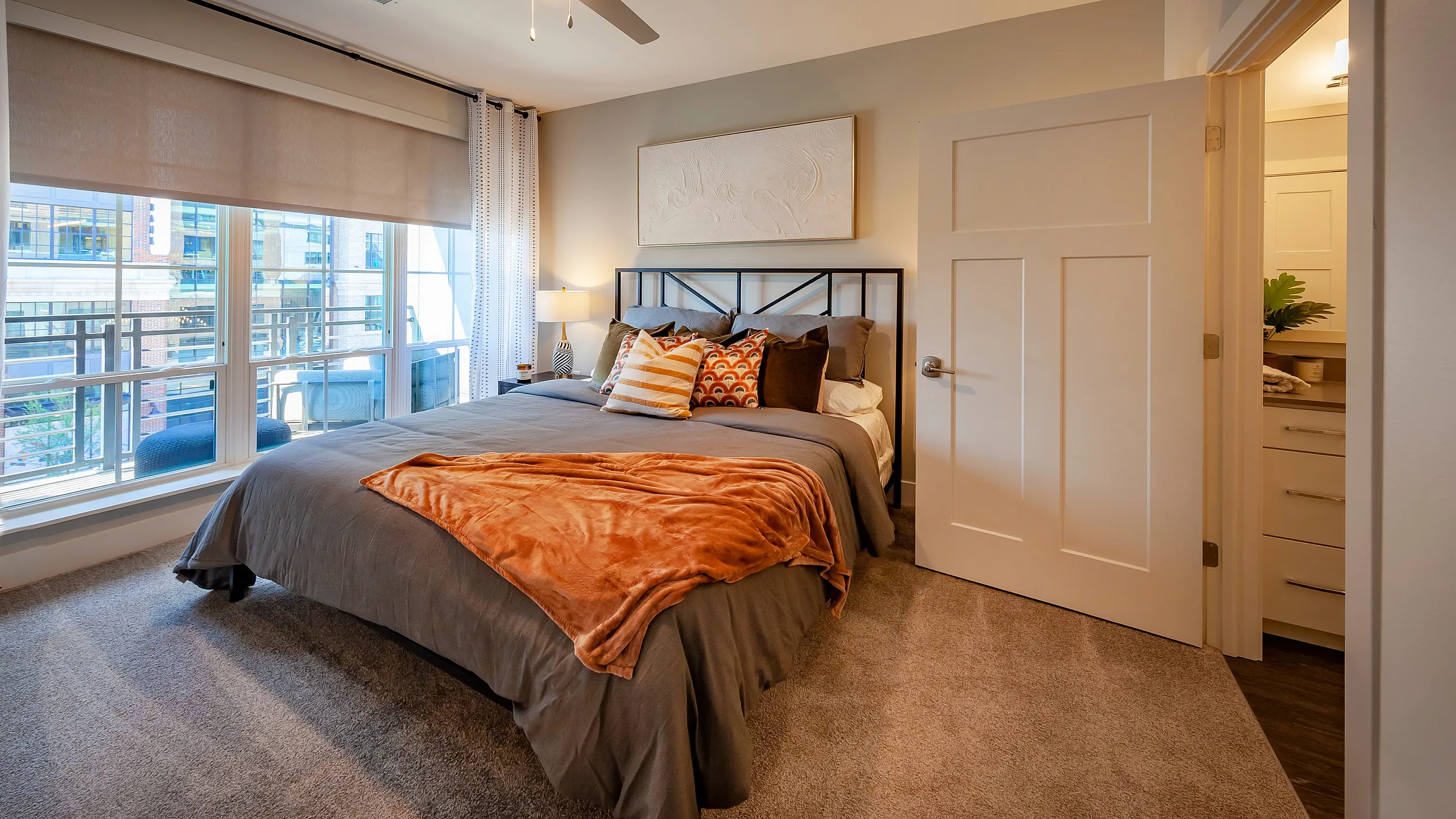 Luxurious bedroom with plush carpet, a stylish bed adorned with decorative pillows, and a warm, inviting atmosphere. Large windows offer a city view, enhancing the room's natural light.