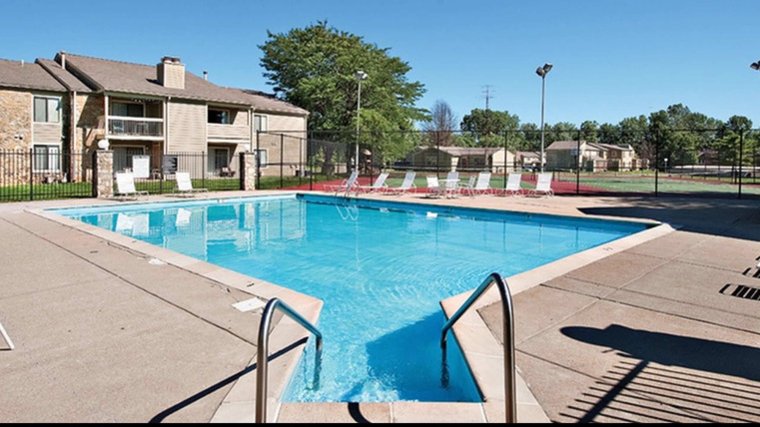 Inviting outdoor pool area with lounge chairs, surrounded by lush greenery and modern apartment buildings under a clear blue sky.