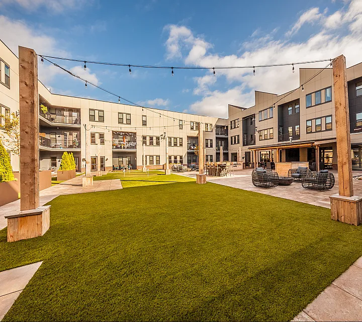Modern apartment complex courtyard with vibrant green lawn, stylish seating, and string lighting under a bright blue sky.