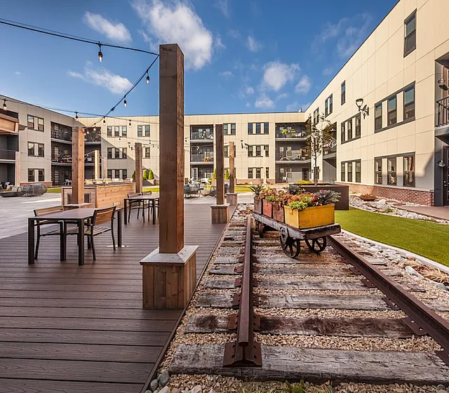 Modern apartment courtyard with charming vintage railway decor, stylish seating areas, and vibrant landscaping under a clear blue sky.