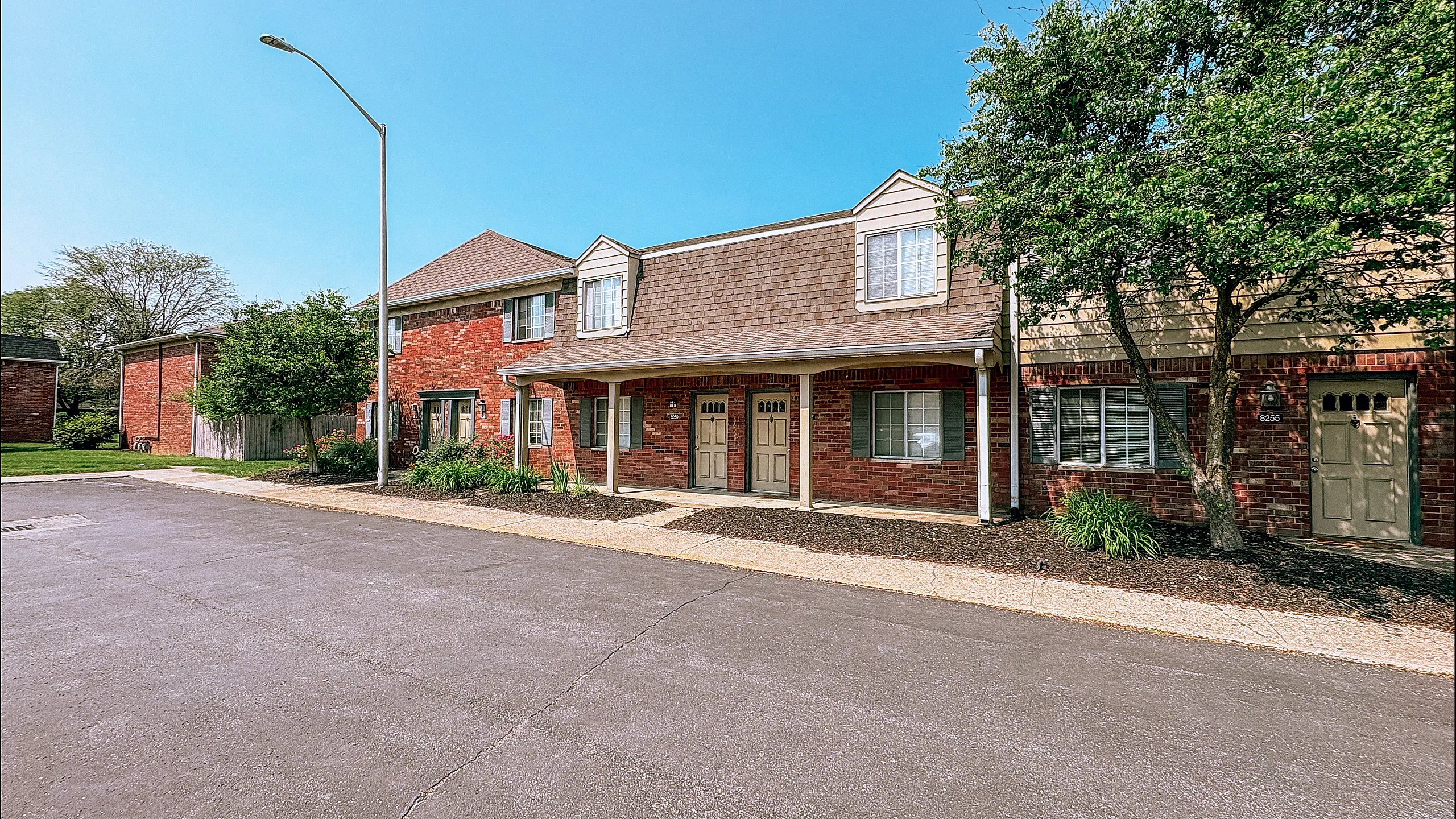 Charming brick apartment complex with lush landscaping, ample natural light, and inviting entrance under a clear blue sky.