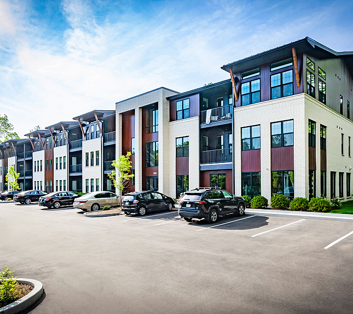 Modern apartment complex with sleek design, large windows, ample parking, and surrounded by lush greenery under a bright blue sky.