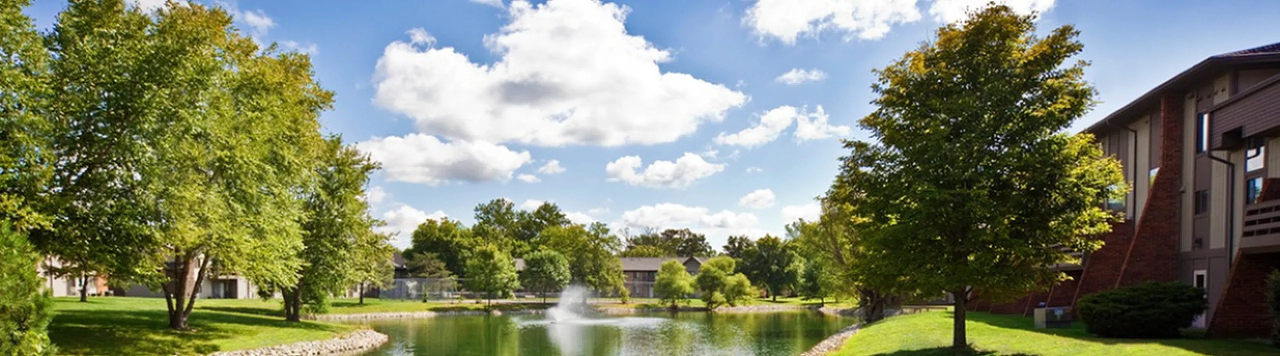 Serene apartment setting with tranquil pond, lush trees, and clear blue skies, perfect for nature lovers.