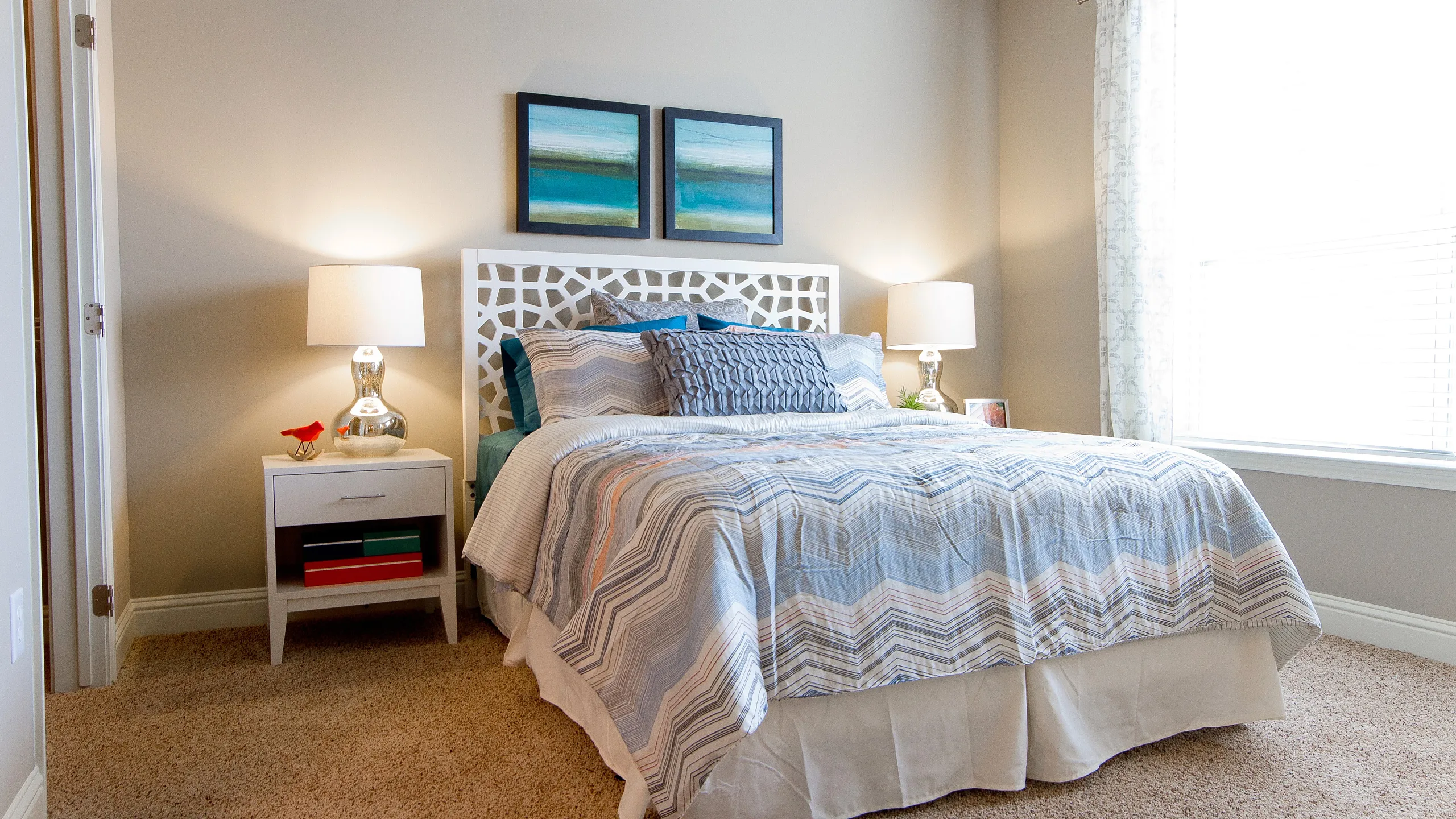 Bright and cozy bedroom with plush carpeting, featuring a stylish bed with patterned bedding, elegant side tables with lamps, and chic wall art.