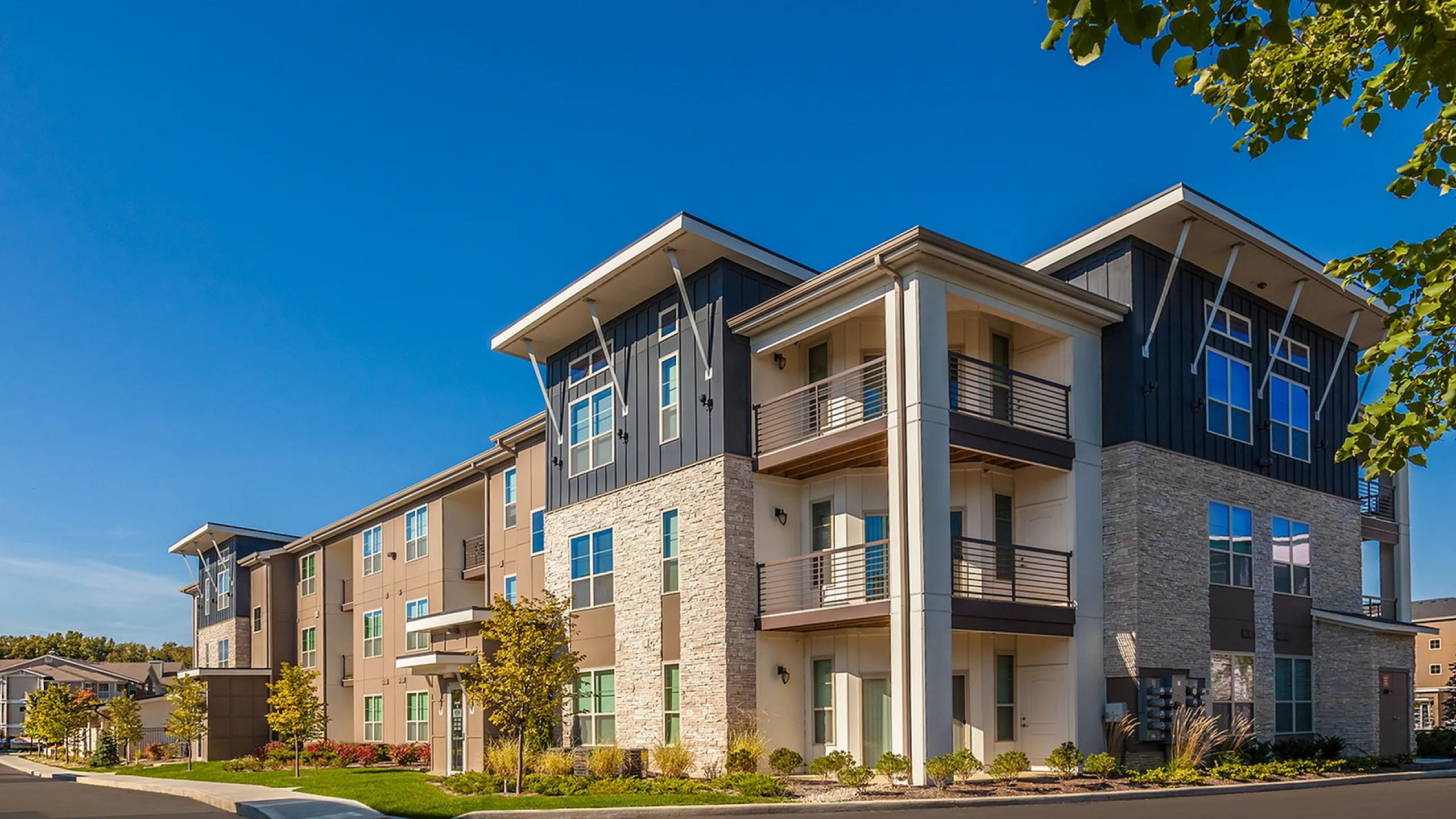 Modern apartment complex exterior with sleek contemporary design, large windows, and welcoming balconies surrounded by lush landscaping under a clear blue sky.
