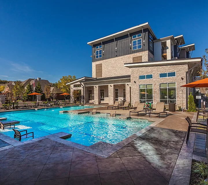 Elegant apartment complex with a modern swimming pool, surrounded by stylish lounge chairs and vibrant orange umbrellas, set against a clear blue sky.