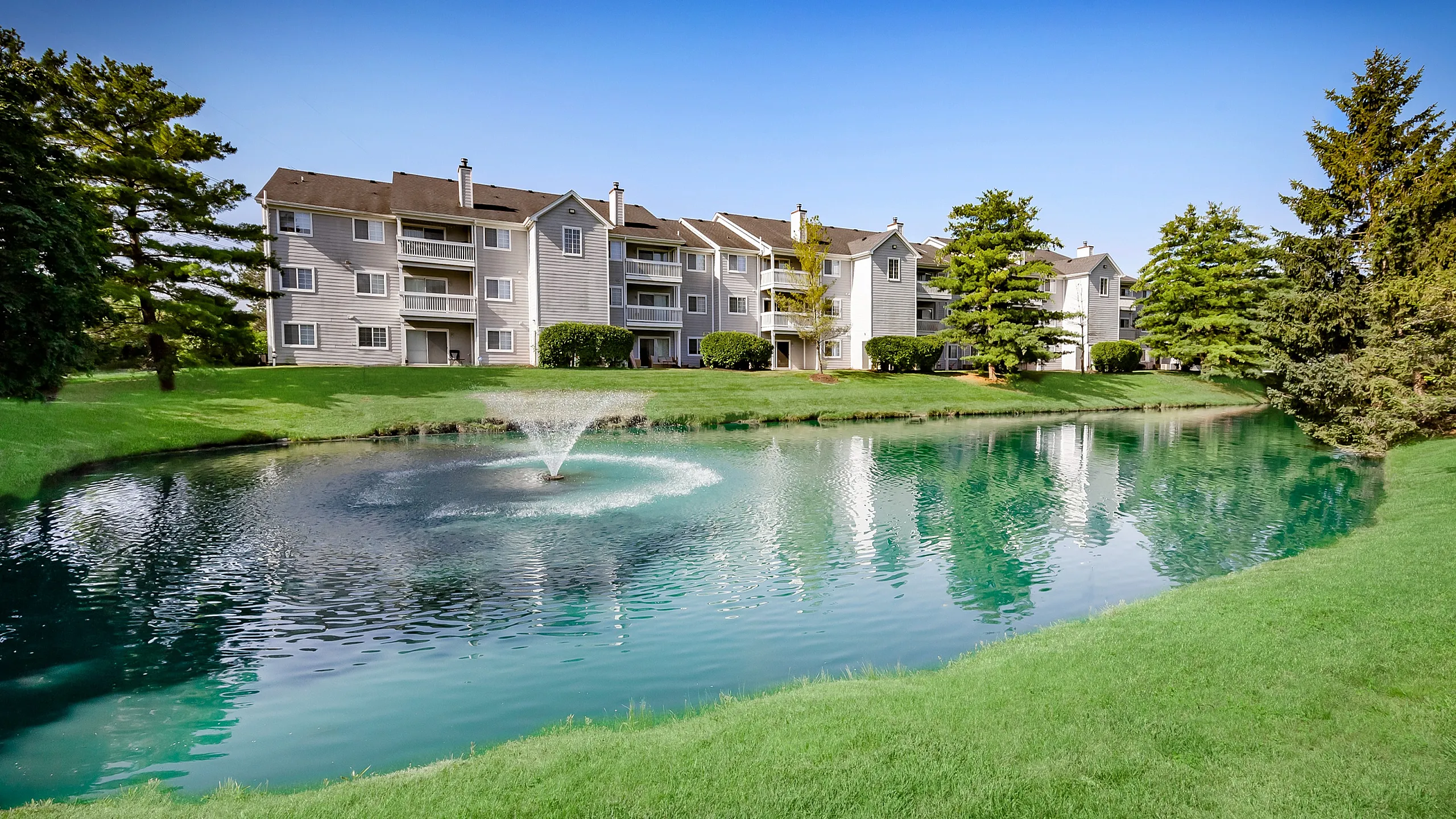 Charming apartment complex showcasing serene water view with a fountain, lush greenery, and inviting, spacious balconies.