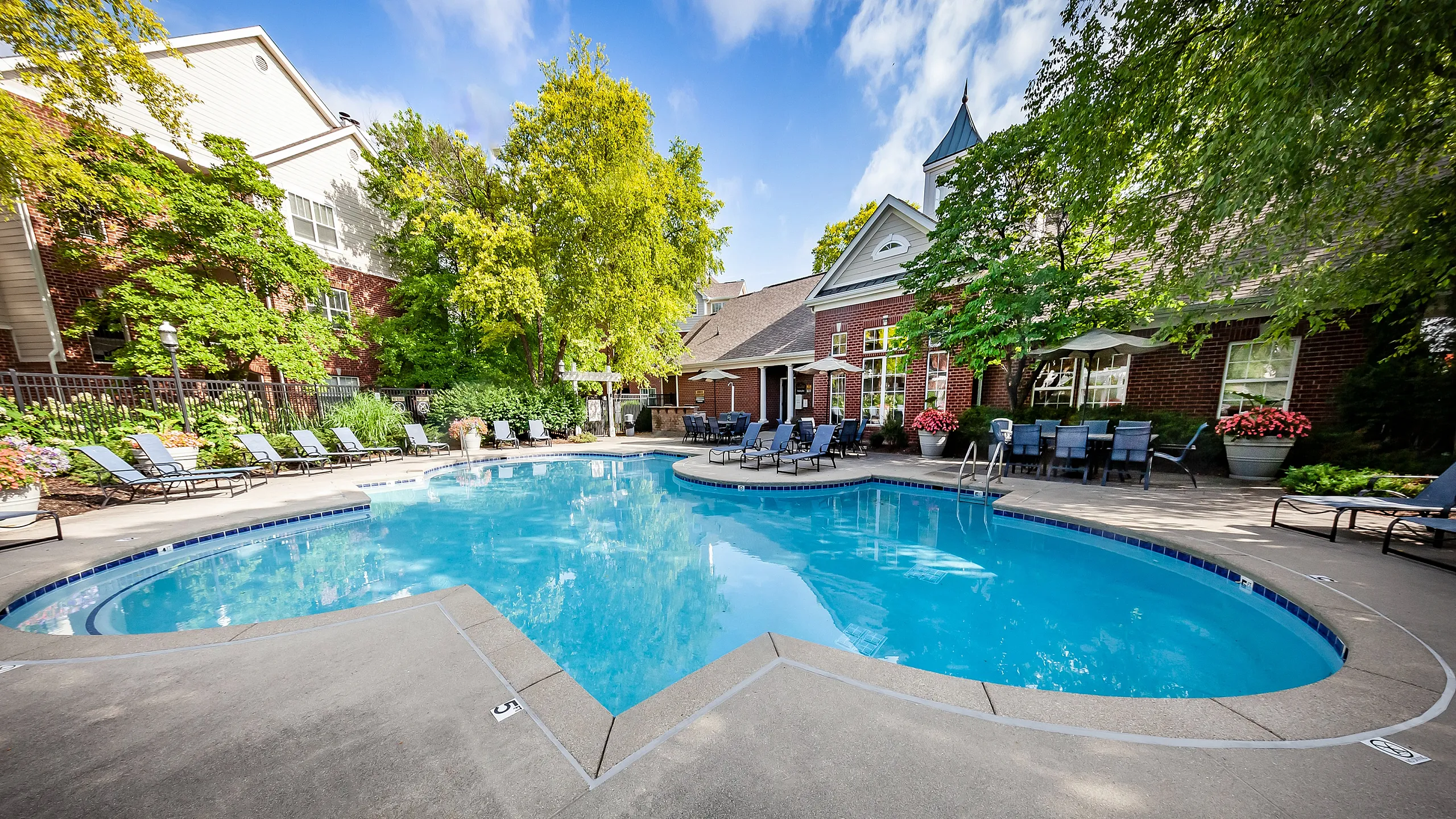 Luxurious outdoor pool area with elegant lounge chairs, surrounded by lush greenery and a charming brick clubhouse.