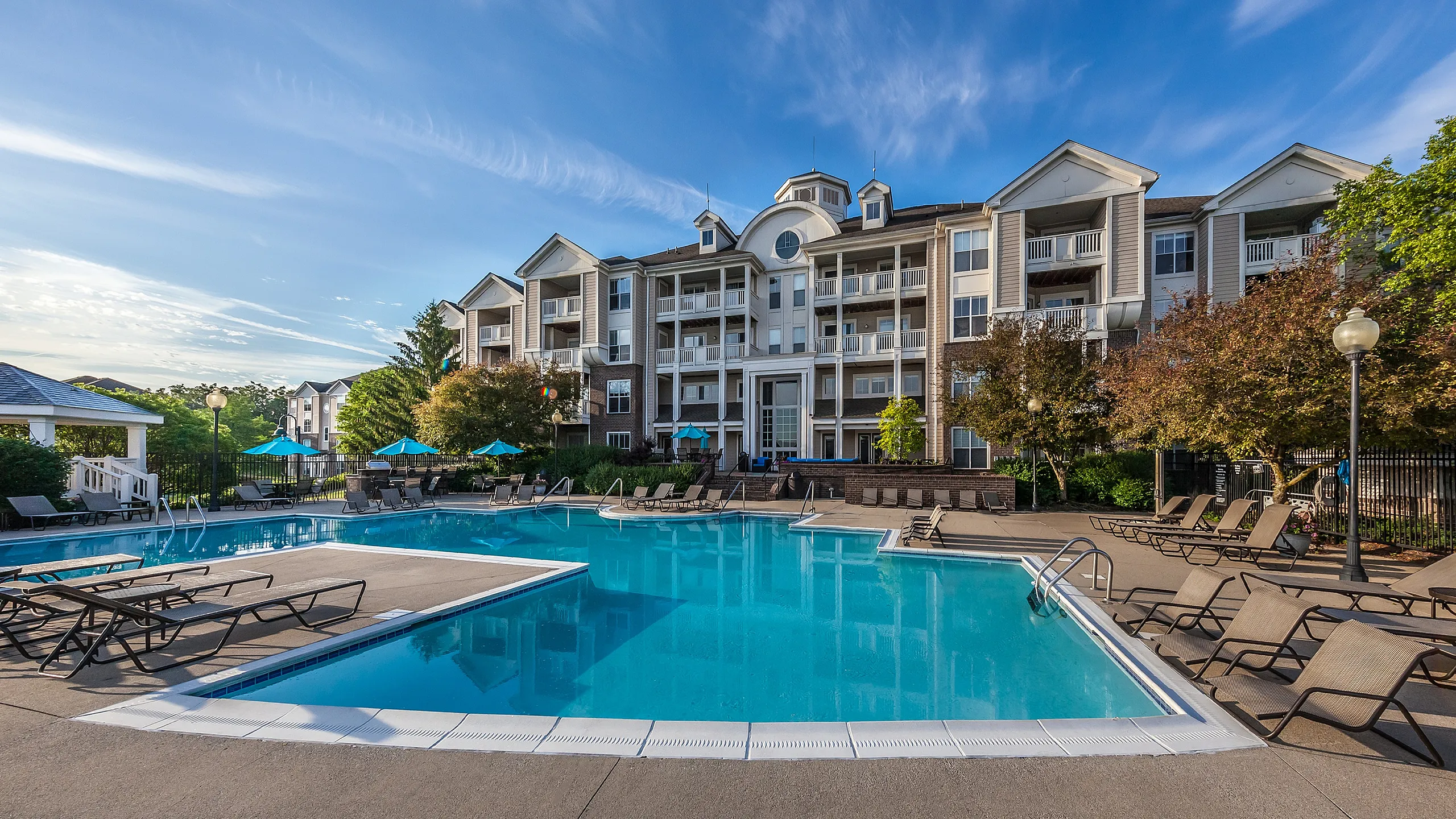 River Crossing Apartments - Luxurious apartment complex with a large outdoor swimming pool, surrounded by stylish lounge chairs and vibrant greenery, under a clear blue sky.