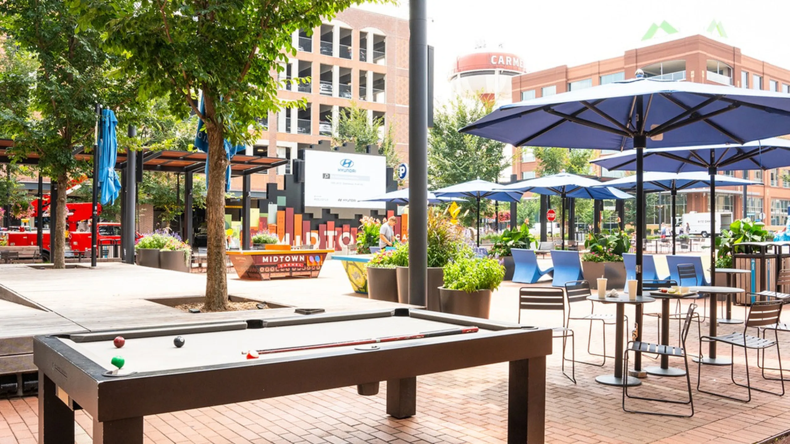 Main Street on the Monon Vibrant outdoor patio featuring a modern billiards table, spacious seating with blue umbrellas, surrounded by lush greenery in a lively urban setting.