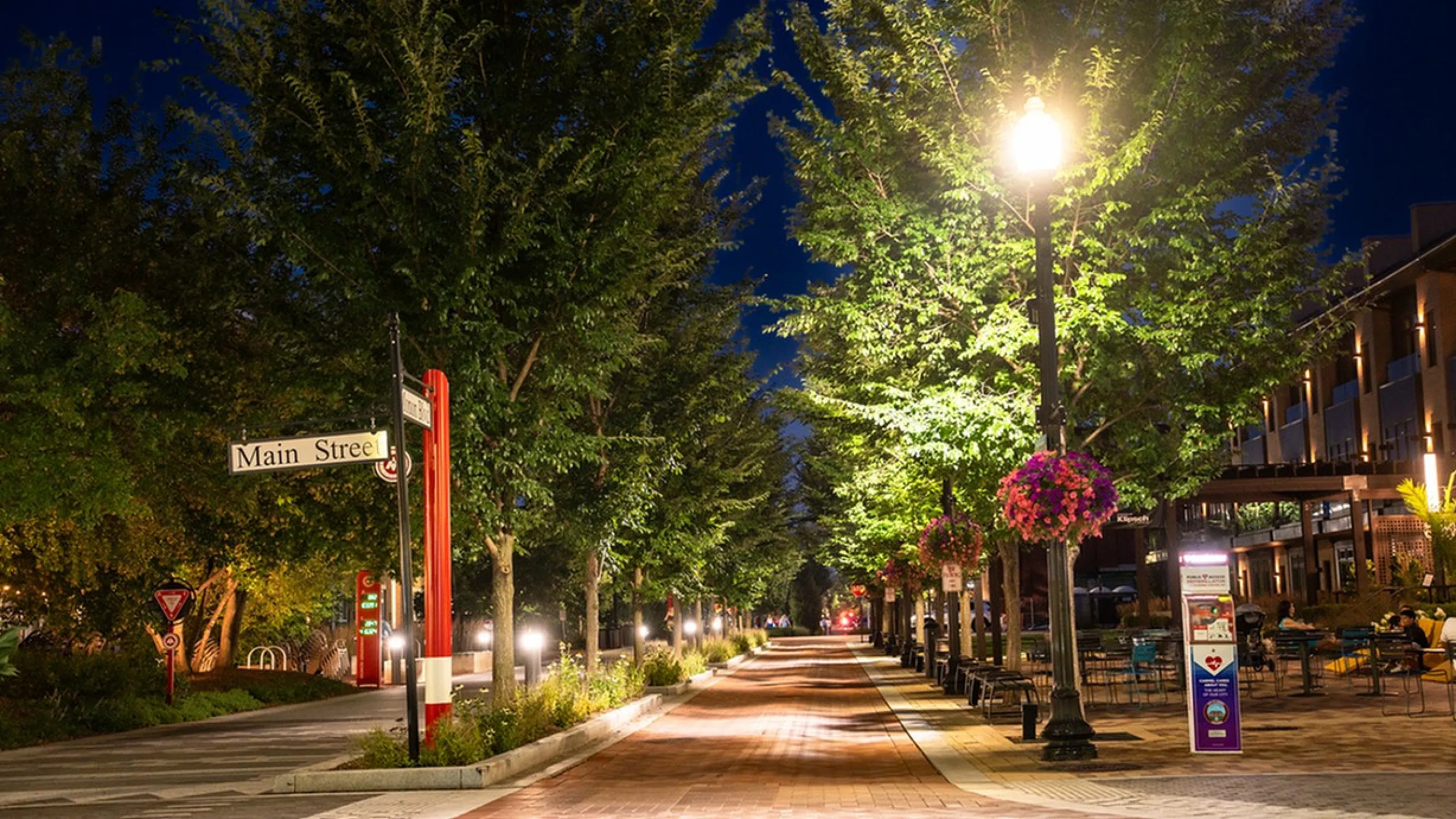 Main Street on the Monon Charming nighttime view of a vibrant Main Street, lined with lush trees and cozy outdoor seating areas, illuminated by elegant street lamps.