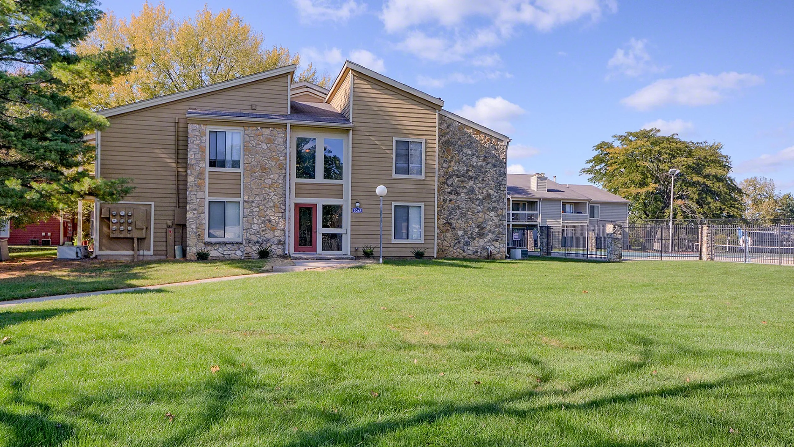 Charming apartment building with rustic stone and wood design, surrounded by lush green lawns and vibrant trees under a clear blue sky.