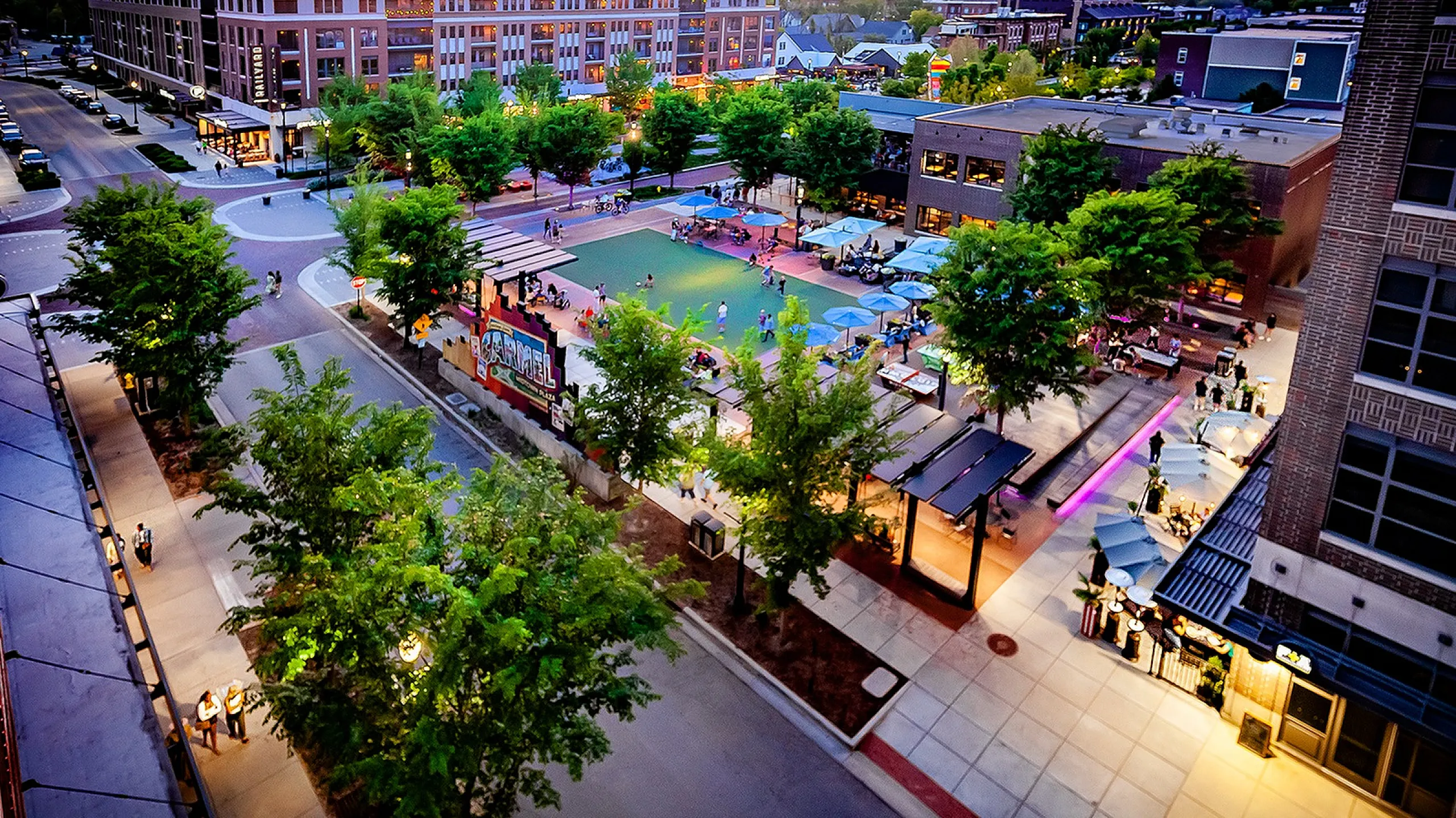 The Railyard at Midtown Apartments - Vibrant urban square at sunset with lively outdoor seating, lush trees, and bustling activity under glowing lights.