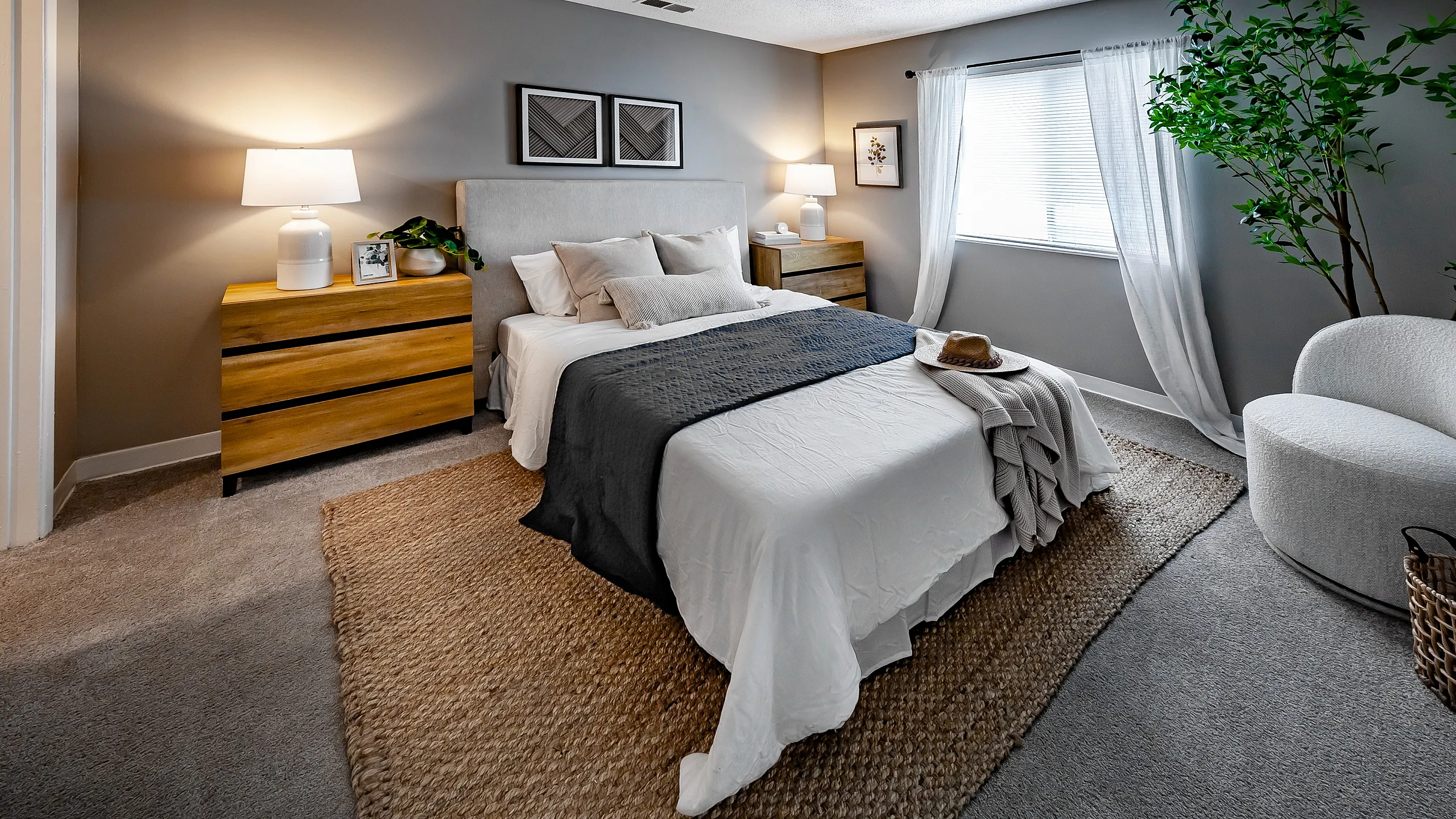 Elegant bedroom featuring a plush bed with layered bedding, flanked by warm wooden dressers, and accented with modern decor and a large leafy plant for a serene ambiance.