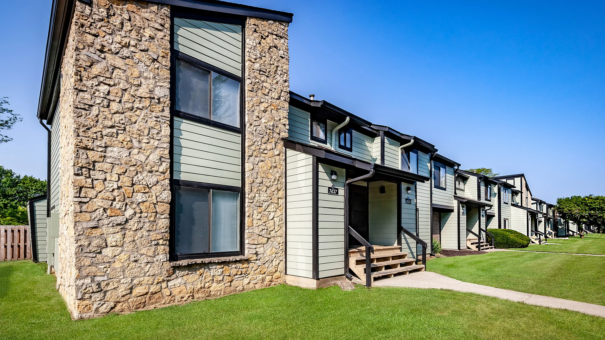 Charming apartment community with rustic stone accents, featuring well-maintained lawns and inviting entrances, set against a clear blue sky.